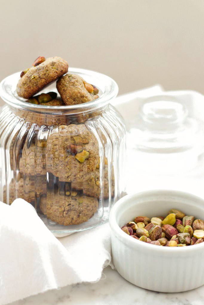 A jar of pistachio cookies with a plate of raw pistachio nuts.