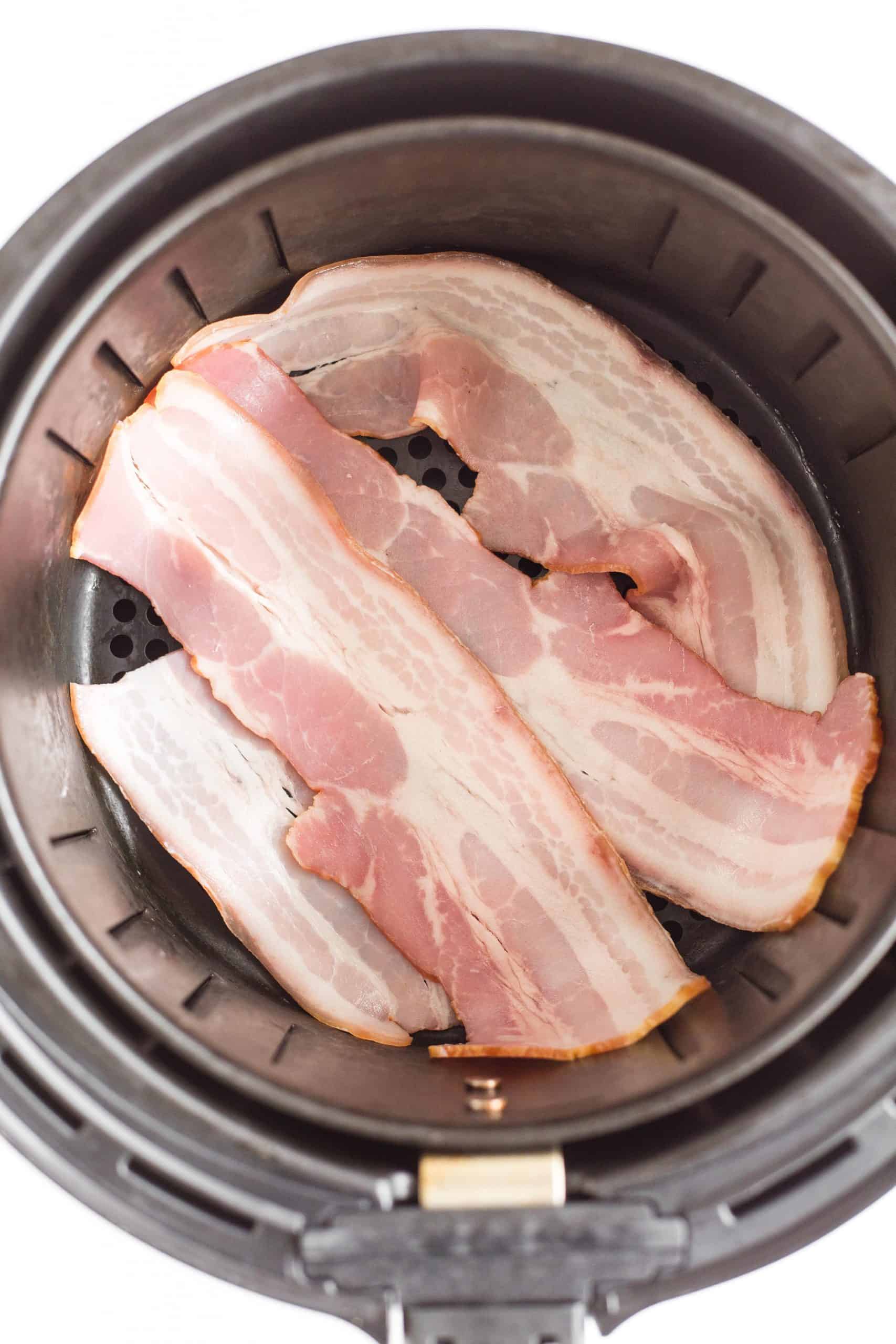 Uncooked bacon slices in an air fryer basket.