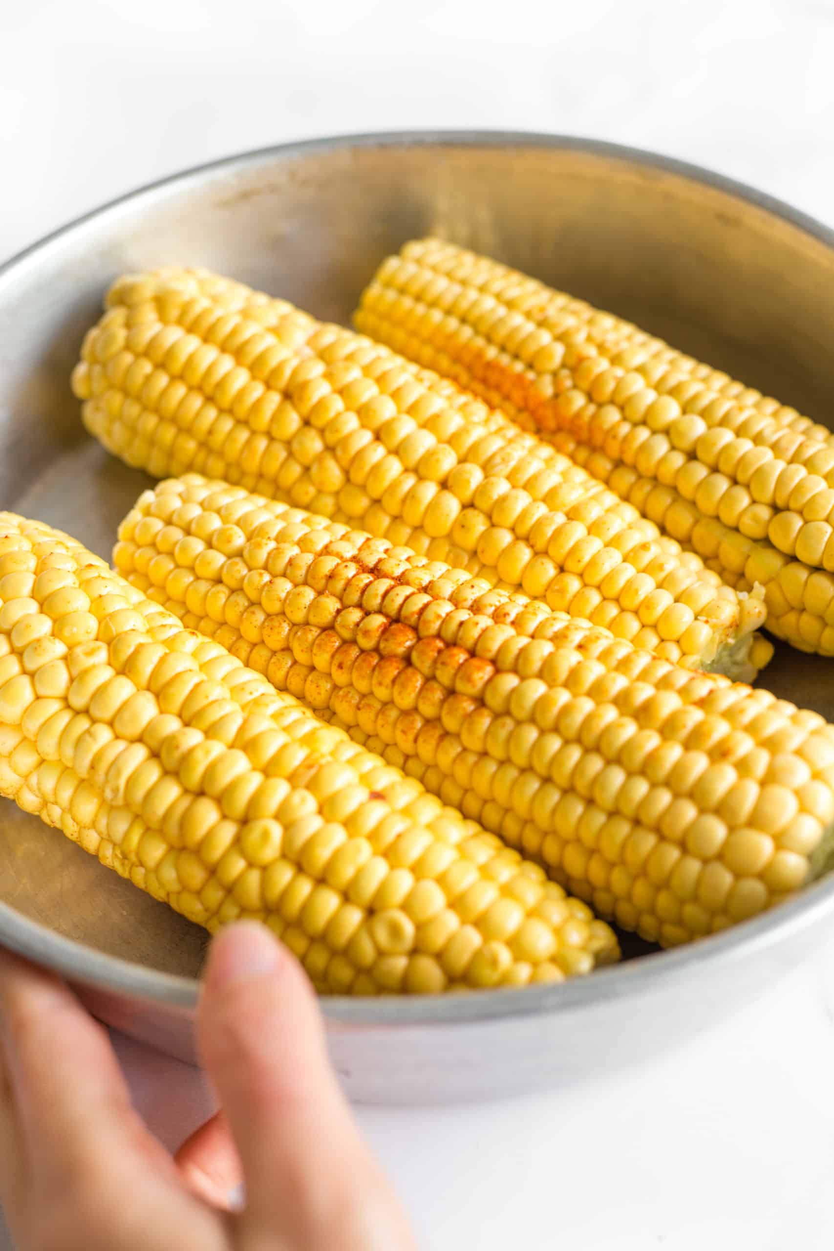 Hand holding a metal bowl of fresh corn on the cob sprinkled with ground paprika.