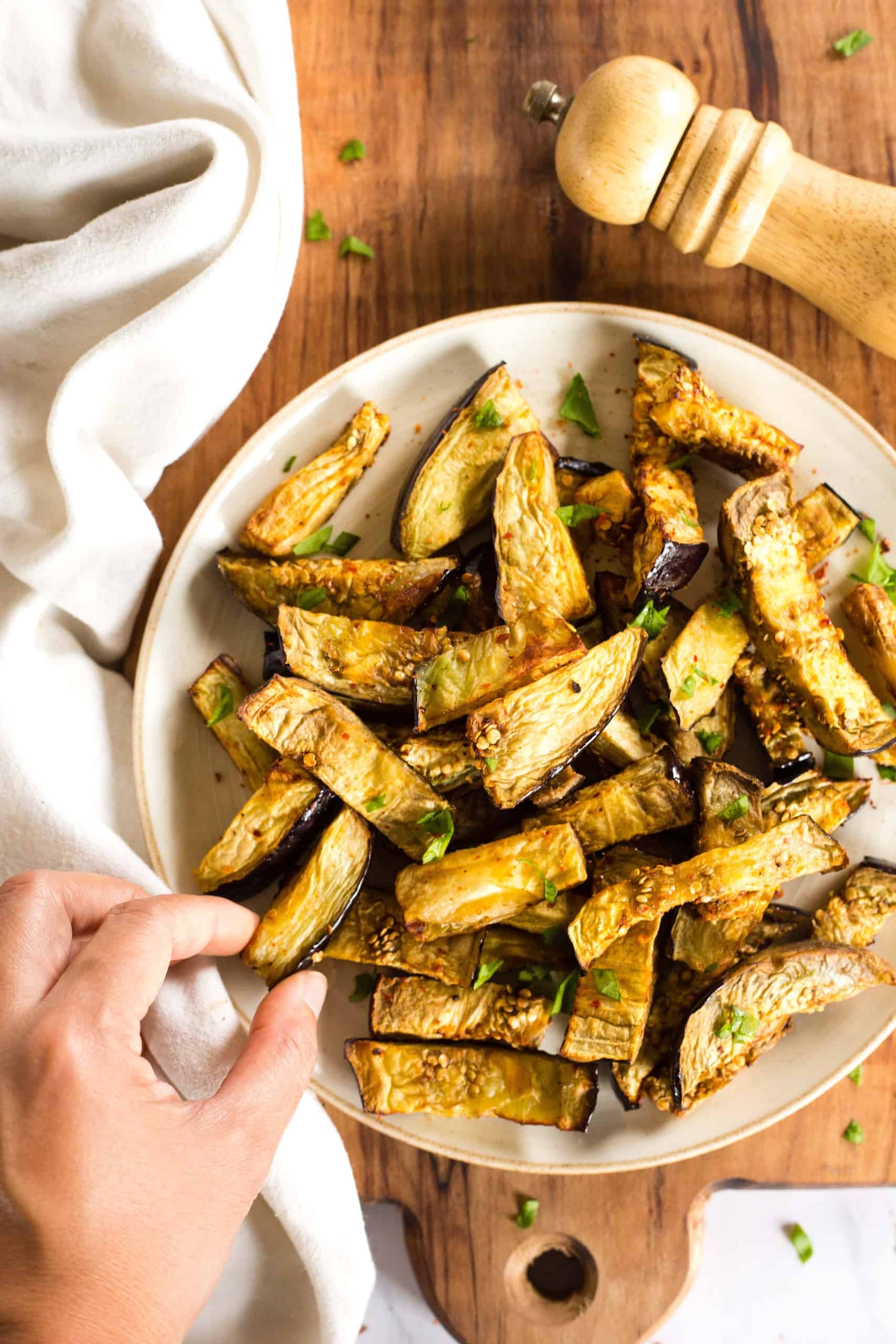 Hand reaching for air fryer eggplant fries from a plate on a wooden board.
