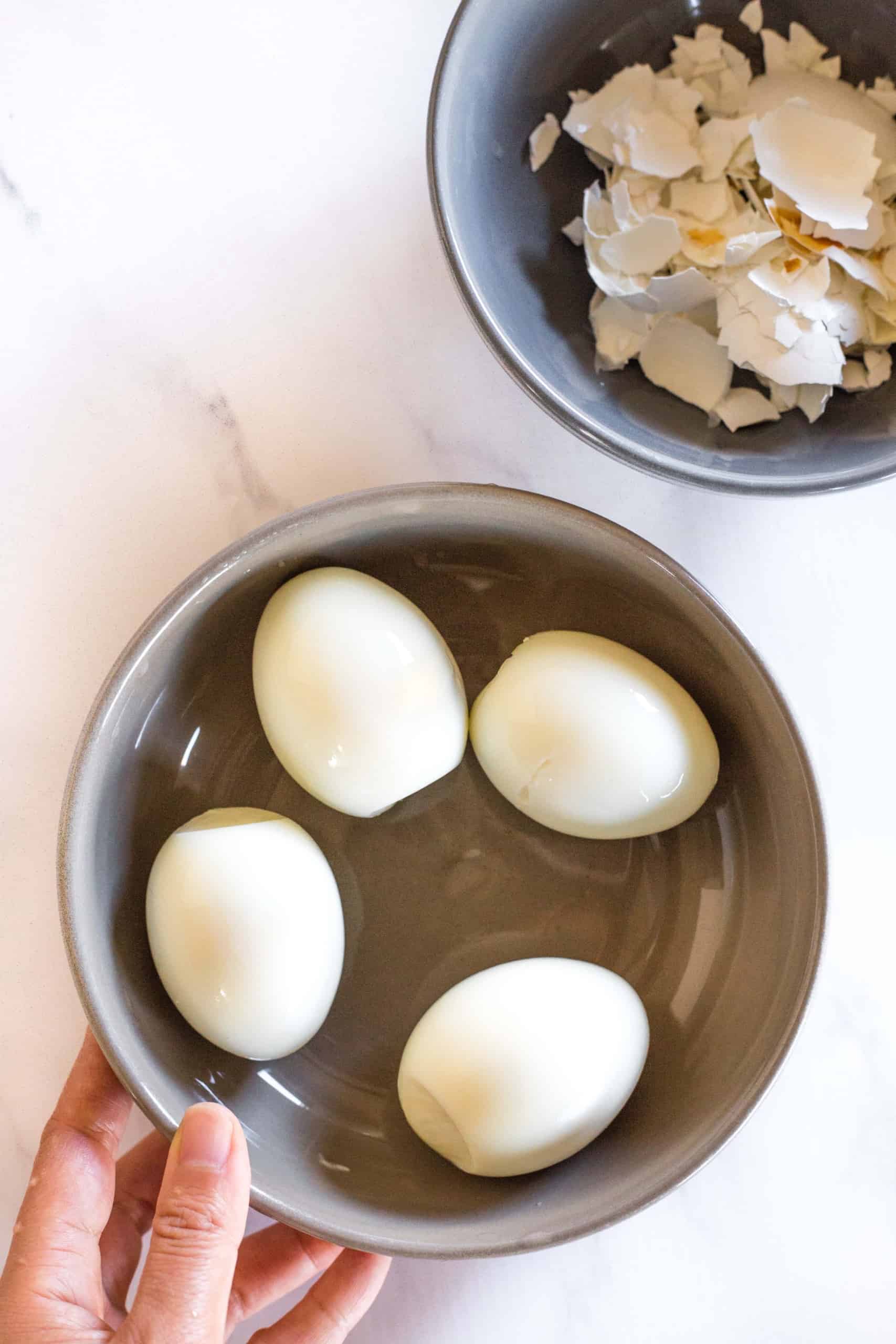 Peeled hard-boiled eggs in a bowl with their shells in another bowl.