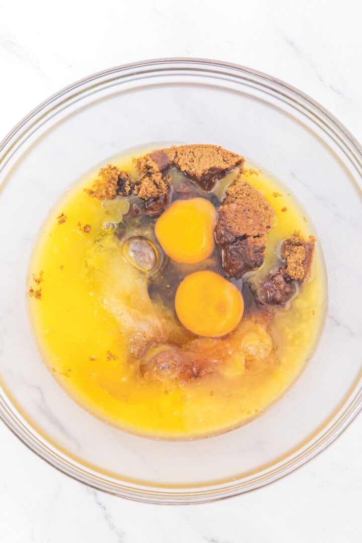 A glass mixing bowl with brown sugar, sugar, melted butter, vanilla extract and eggs.