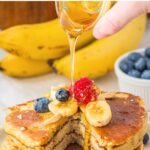 Pouring maple syrup over a stack of pancakes topped with fruits.