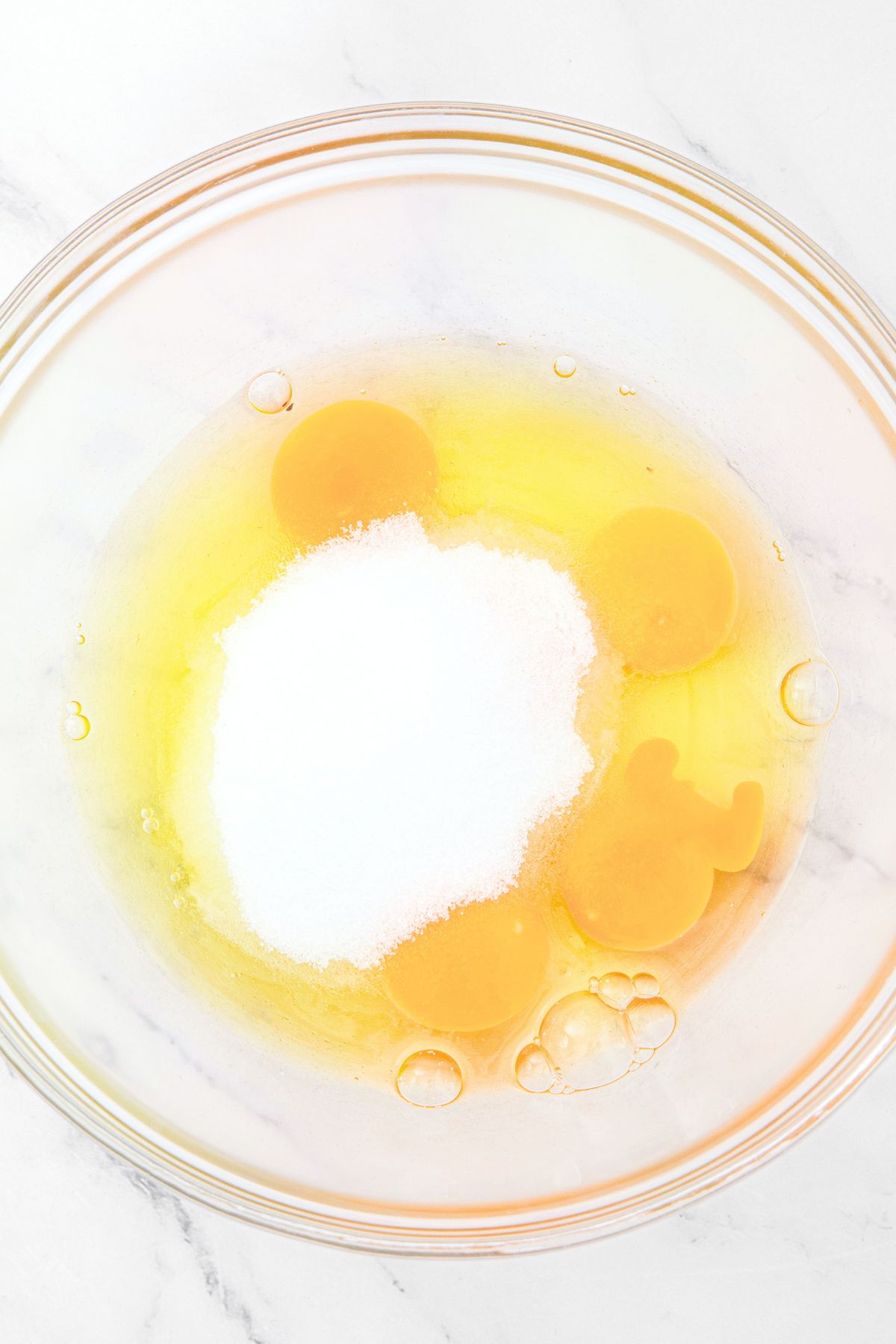 A glass mixing bowl with raw eggs, and sugar.