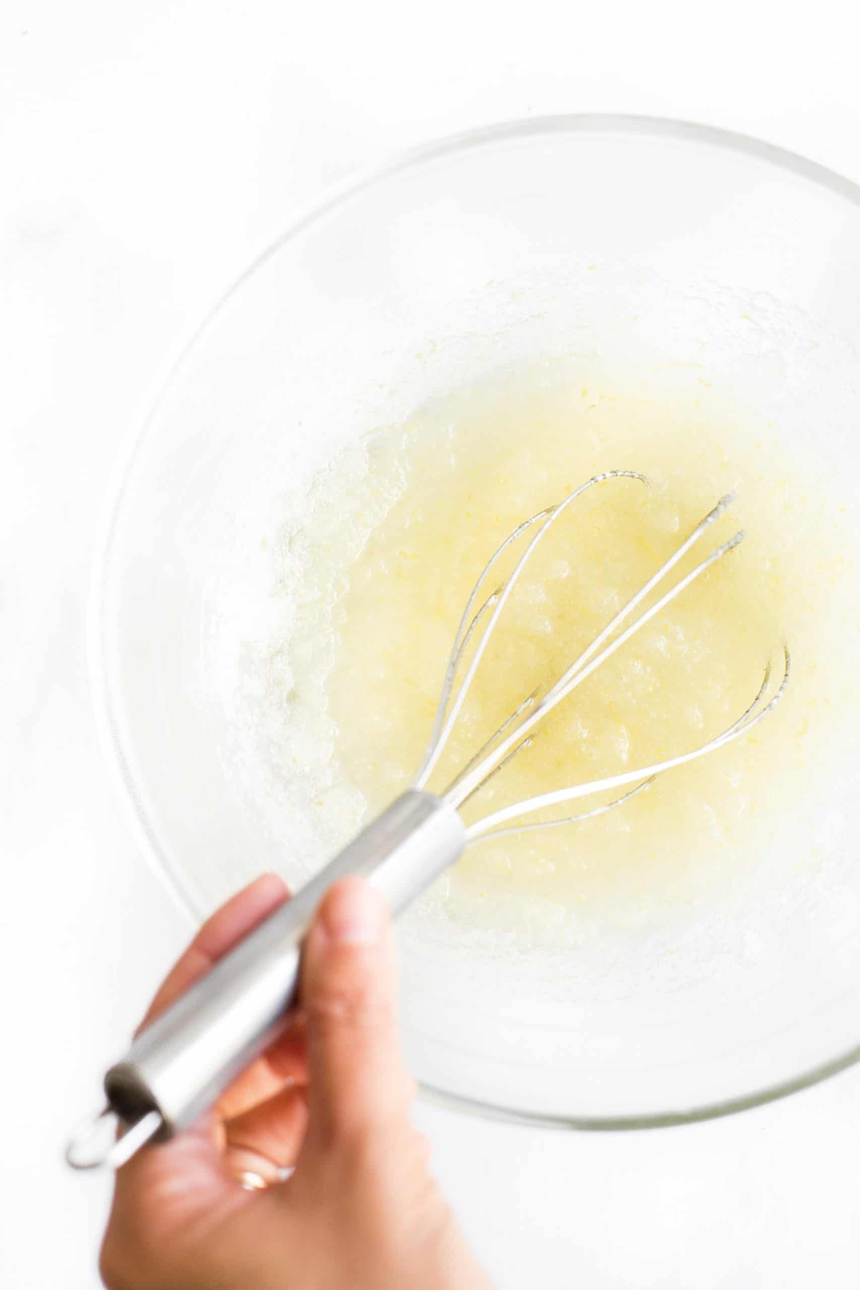 Using a whisk to mix sugar, oil, and zest in a glass bowl.