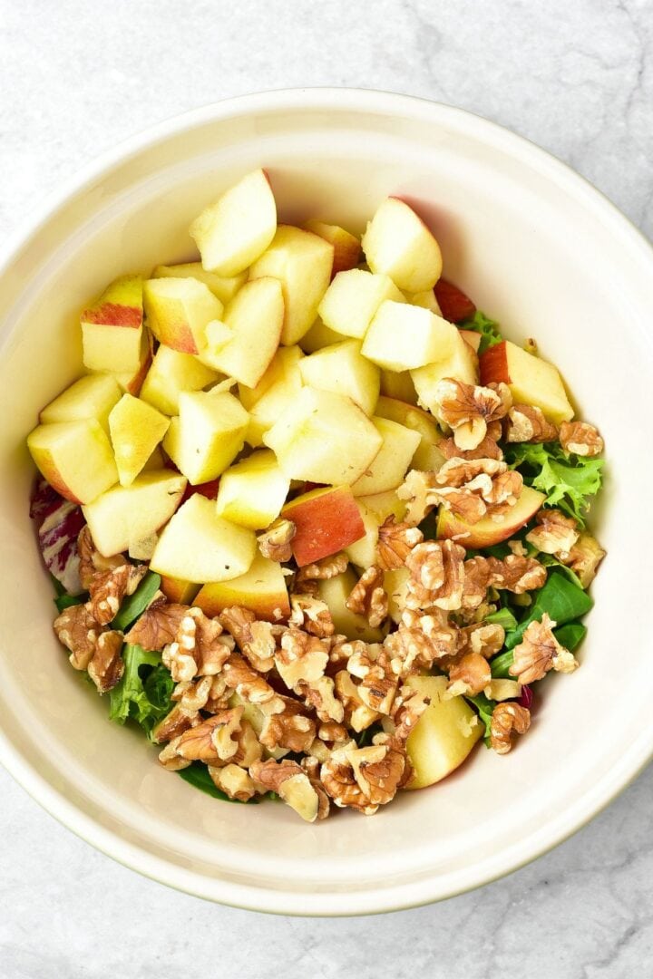 A white bowl with diced apples, chopped walnuts, and salad greens.
