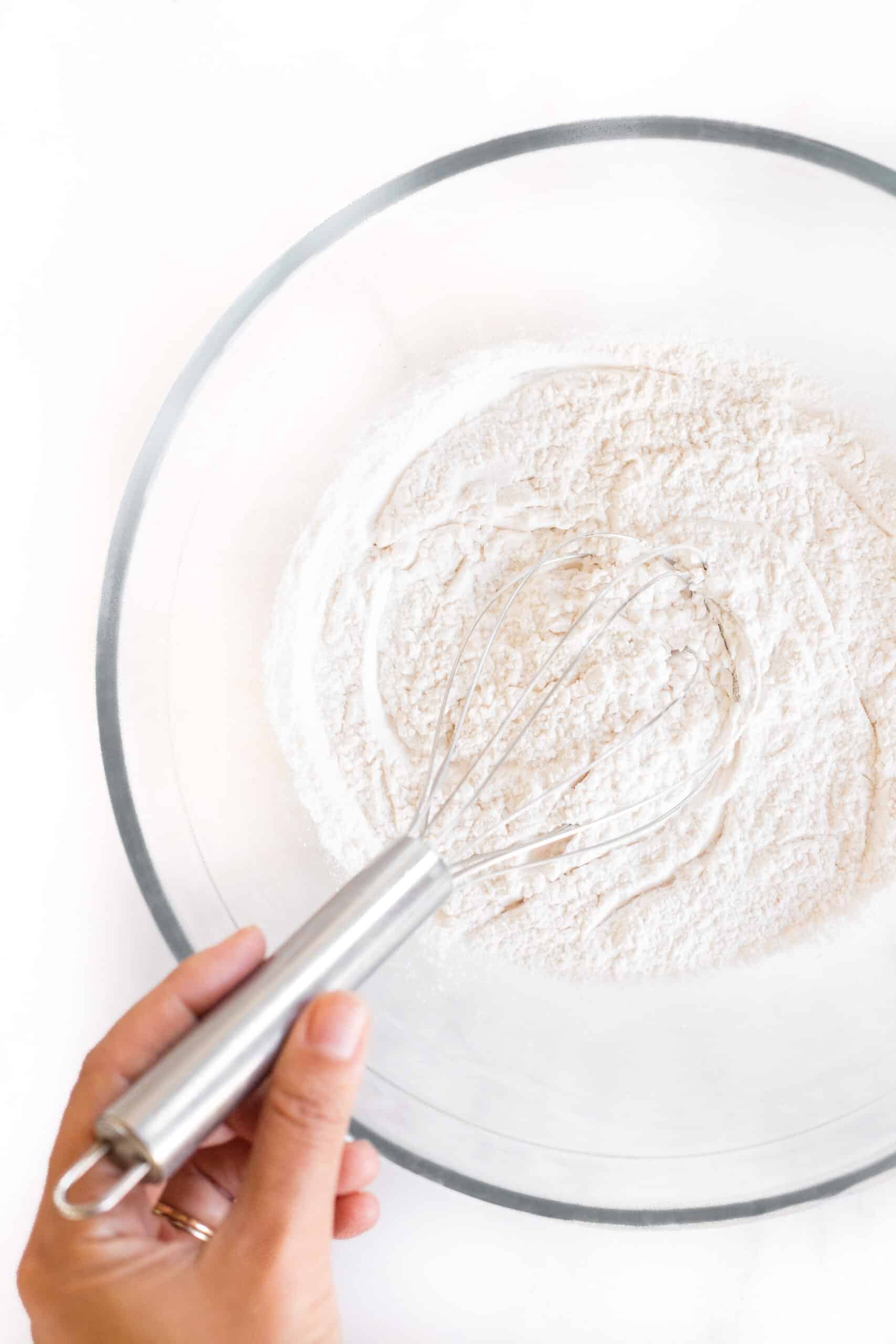 Hand whisking flour mixture in a large glass bowl.