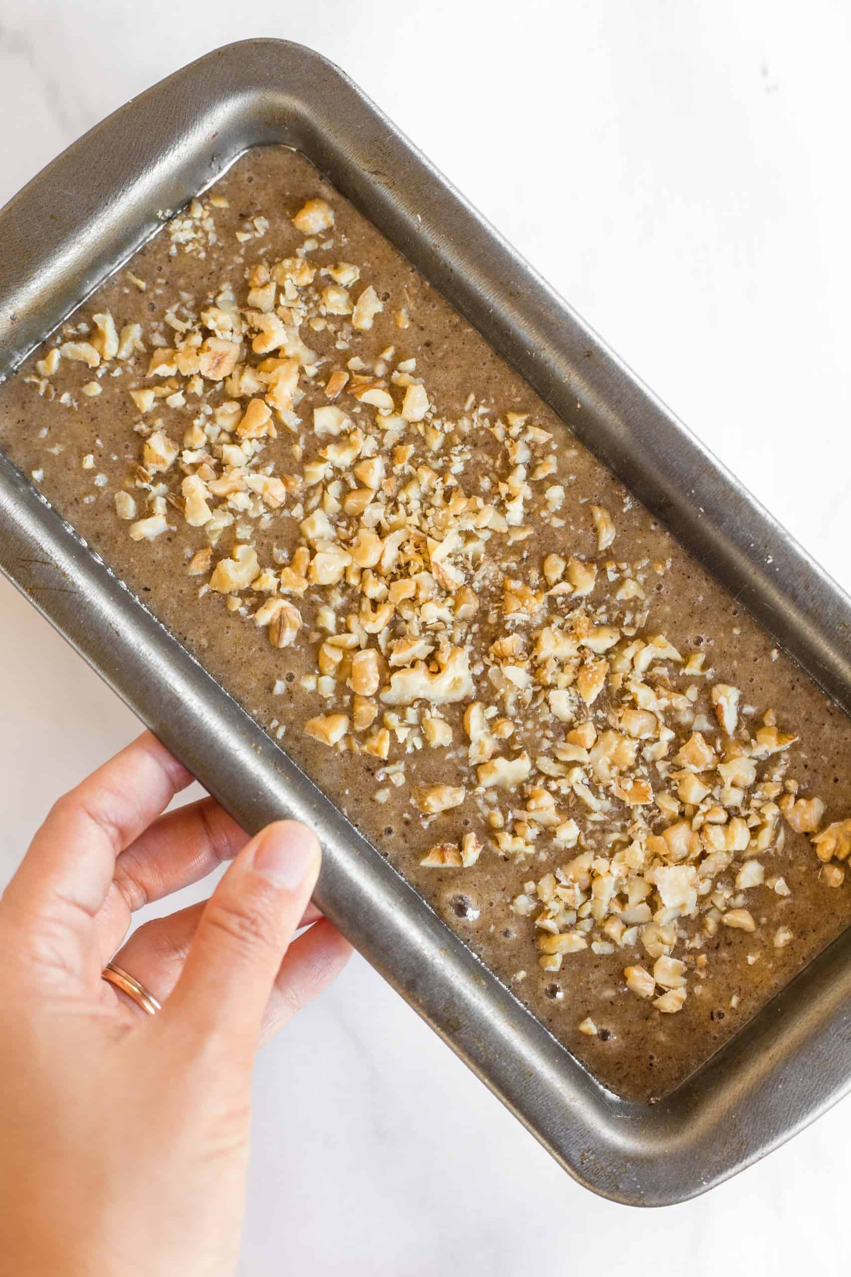 Holding a metal loaf pan with batter sprinkled with chopped walnuts.