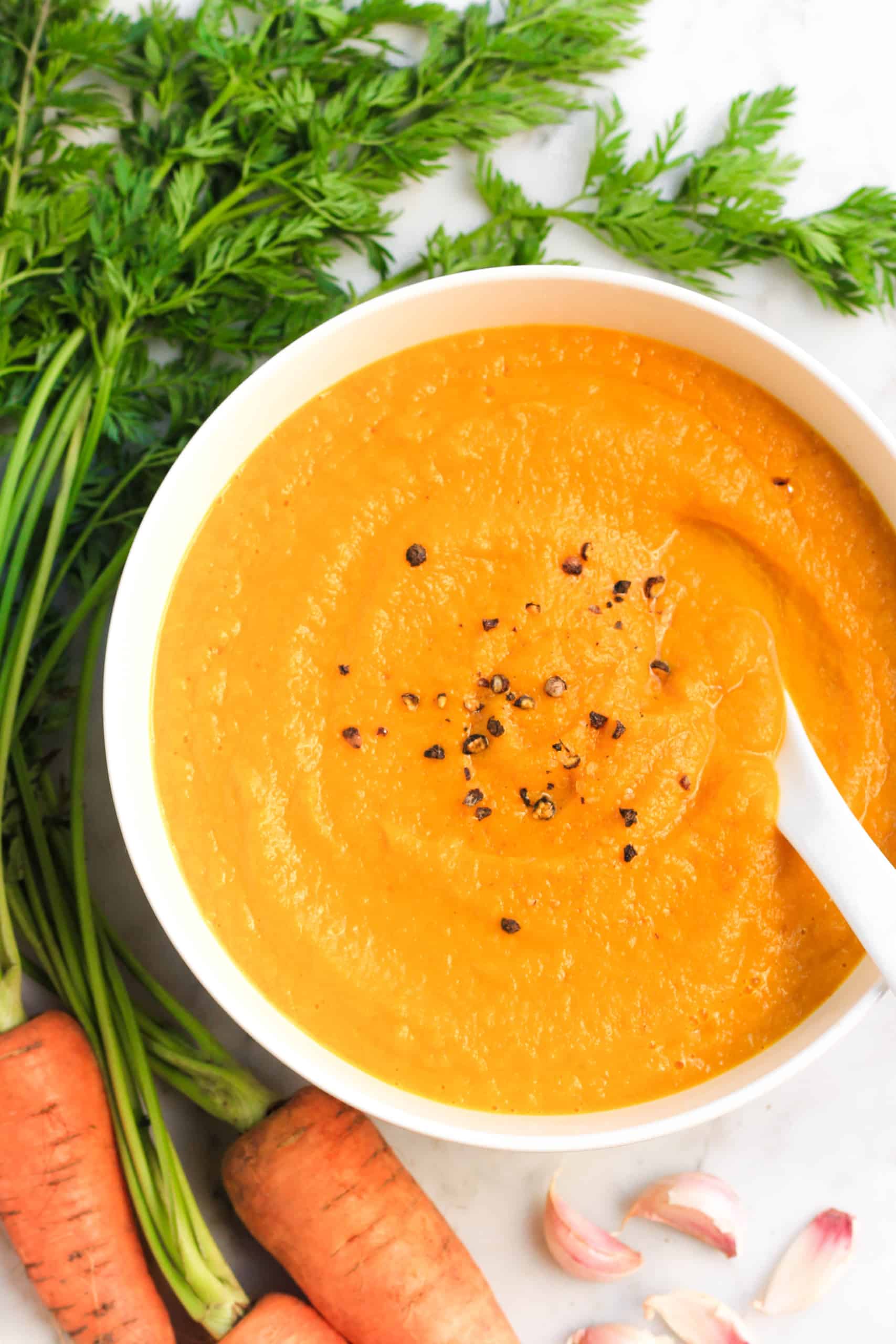 A bowl of cream of carrot soup surrounded by fresh carrots and garlic.