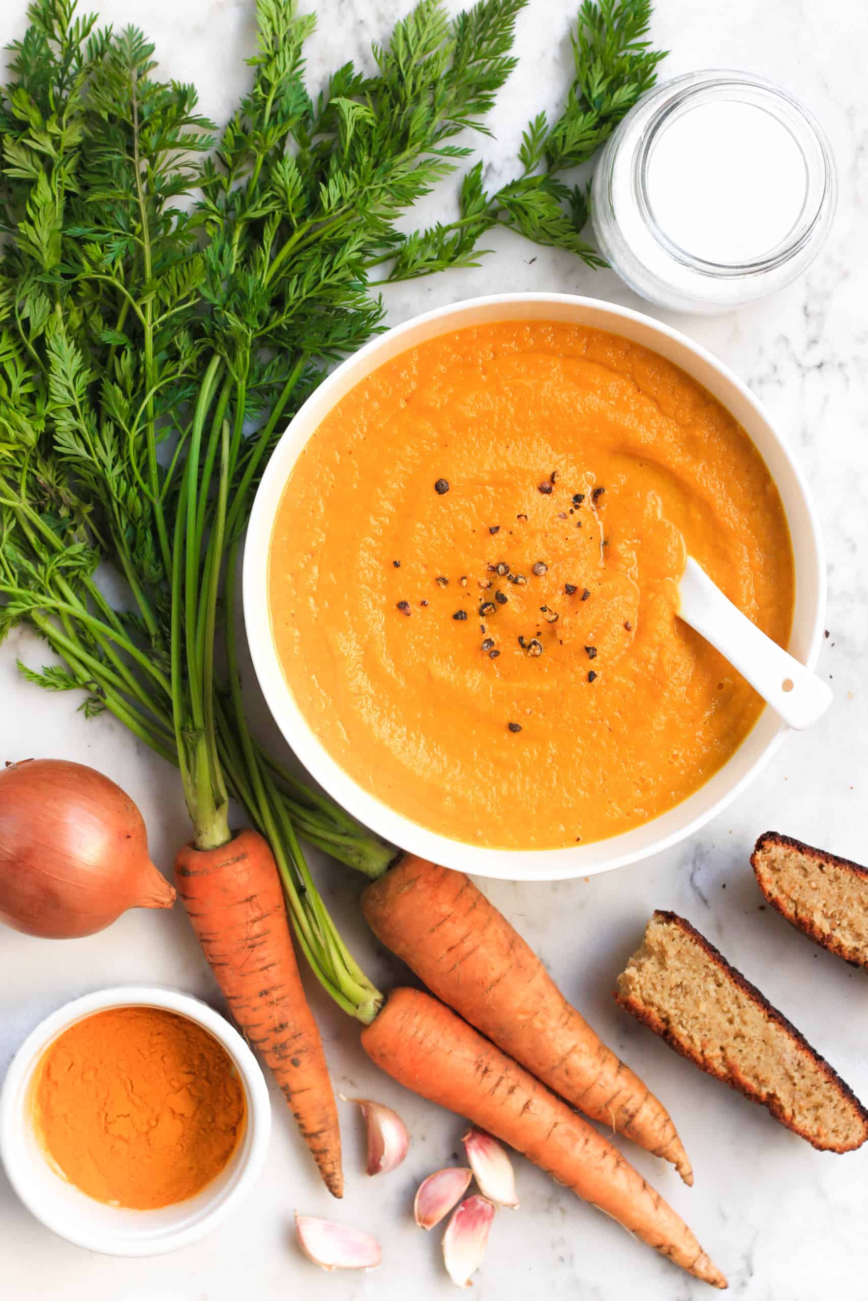 Raw carrots, fresh bread, and a bowl of creamy carrot turmeric soup on marble board.