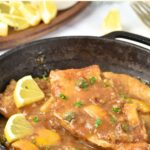 A skillet of chicken piccata and slices of lemon behind.