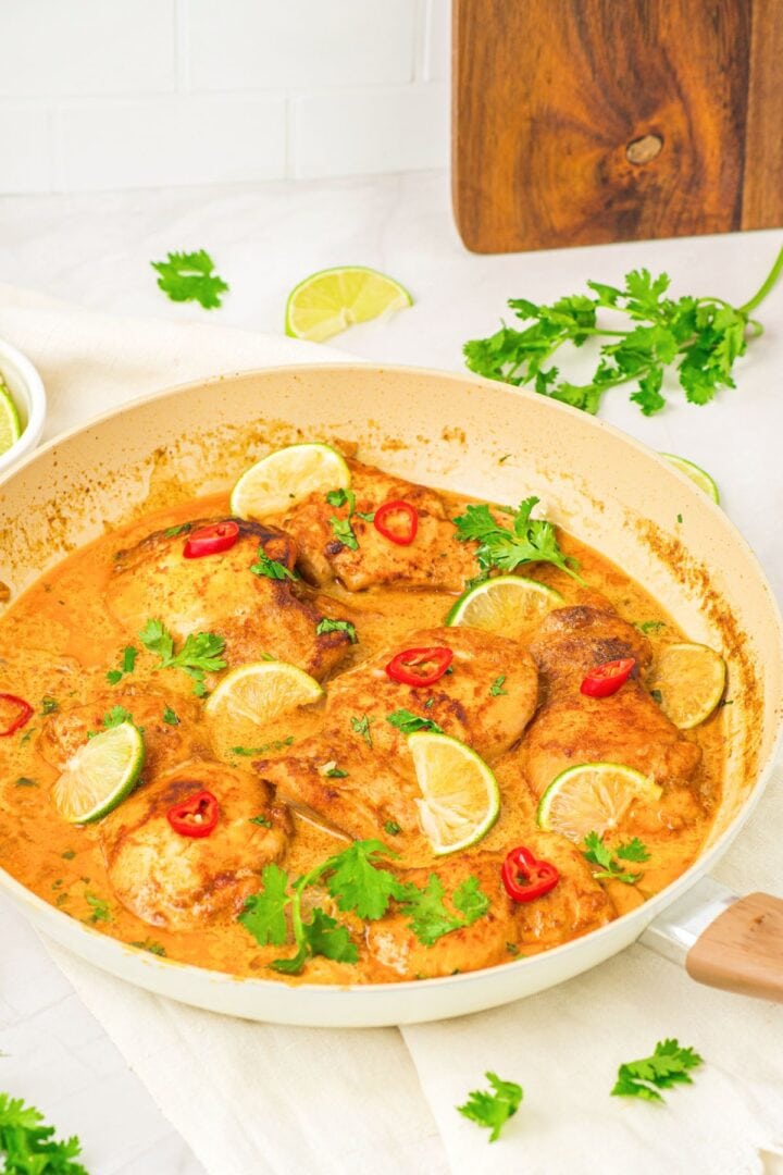 A skillet with coconut lime chicken thighs topped with lime wedges and red chili slices.