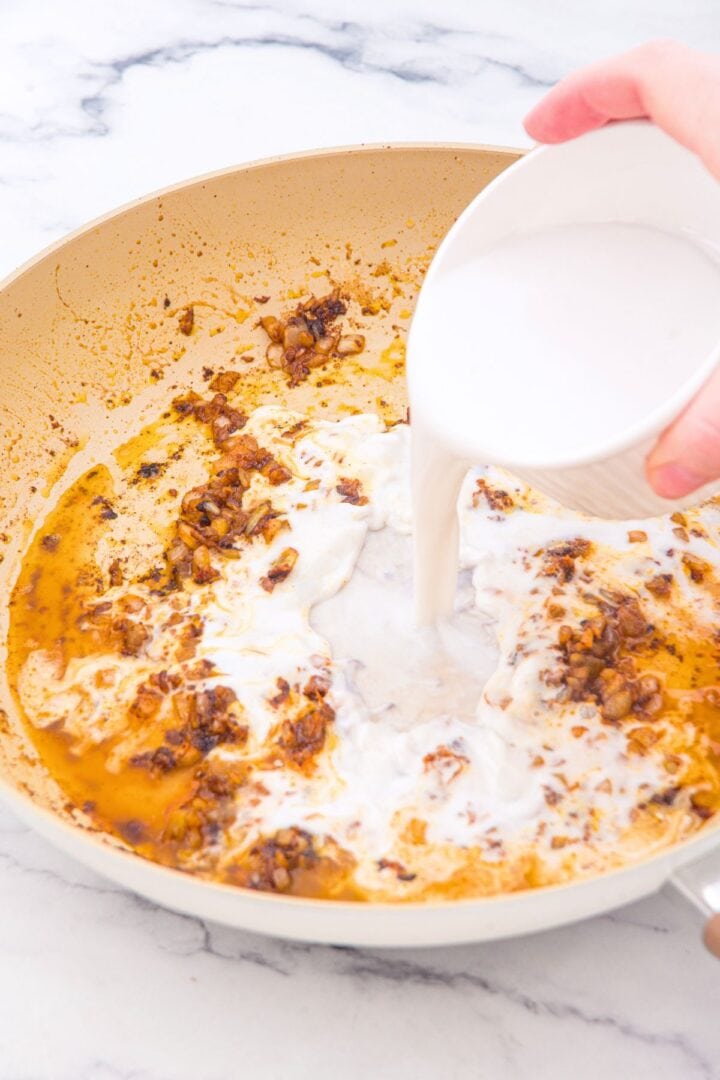 Pouring coconut milk into a skillet with sautéed onions.