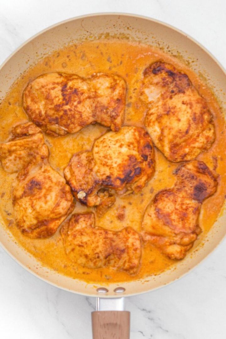 Top down view of chicken thighs simmering in a orange liquid mixture in skillet.