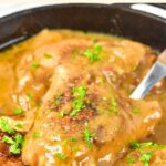 Up close view of a spoon holding up chicken thigh from a skillet