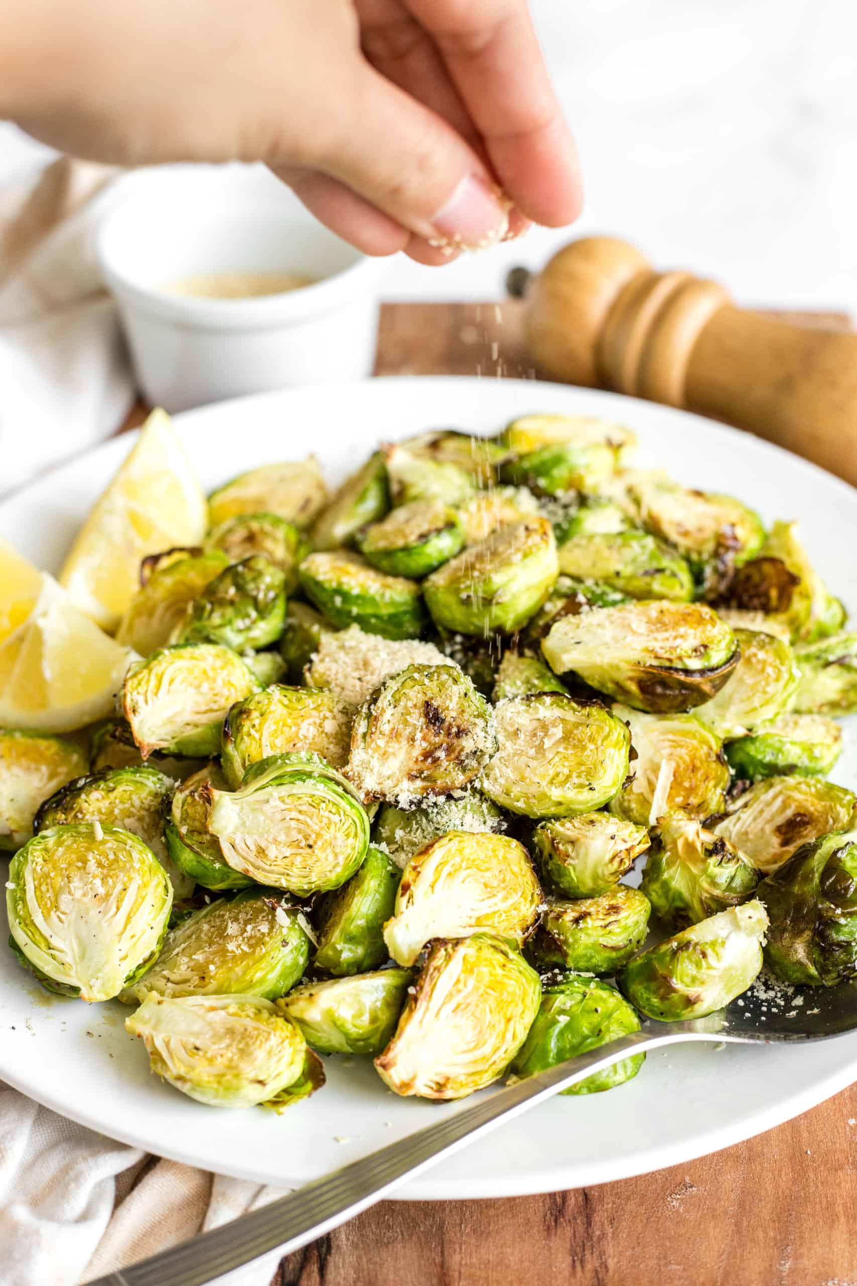 A hand sprinkling nutrional yeast on top of a plate full of crispy Brussels sprouts.