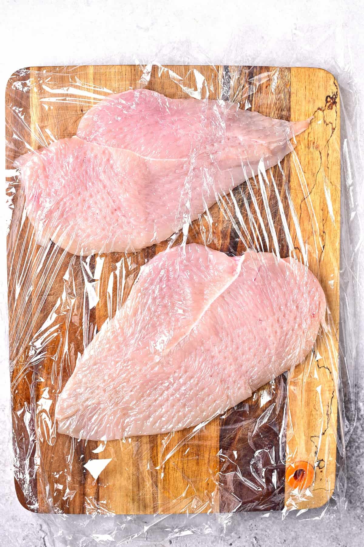 Chicken cutlets on wooden board covered with plastic wrap.