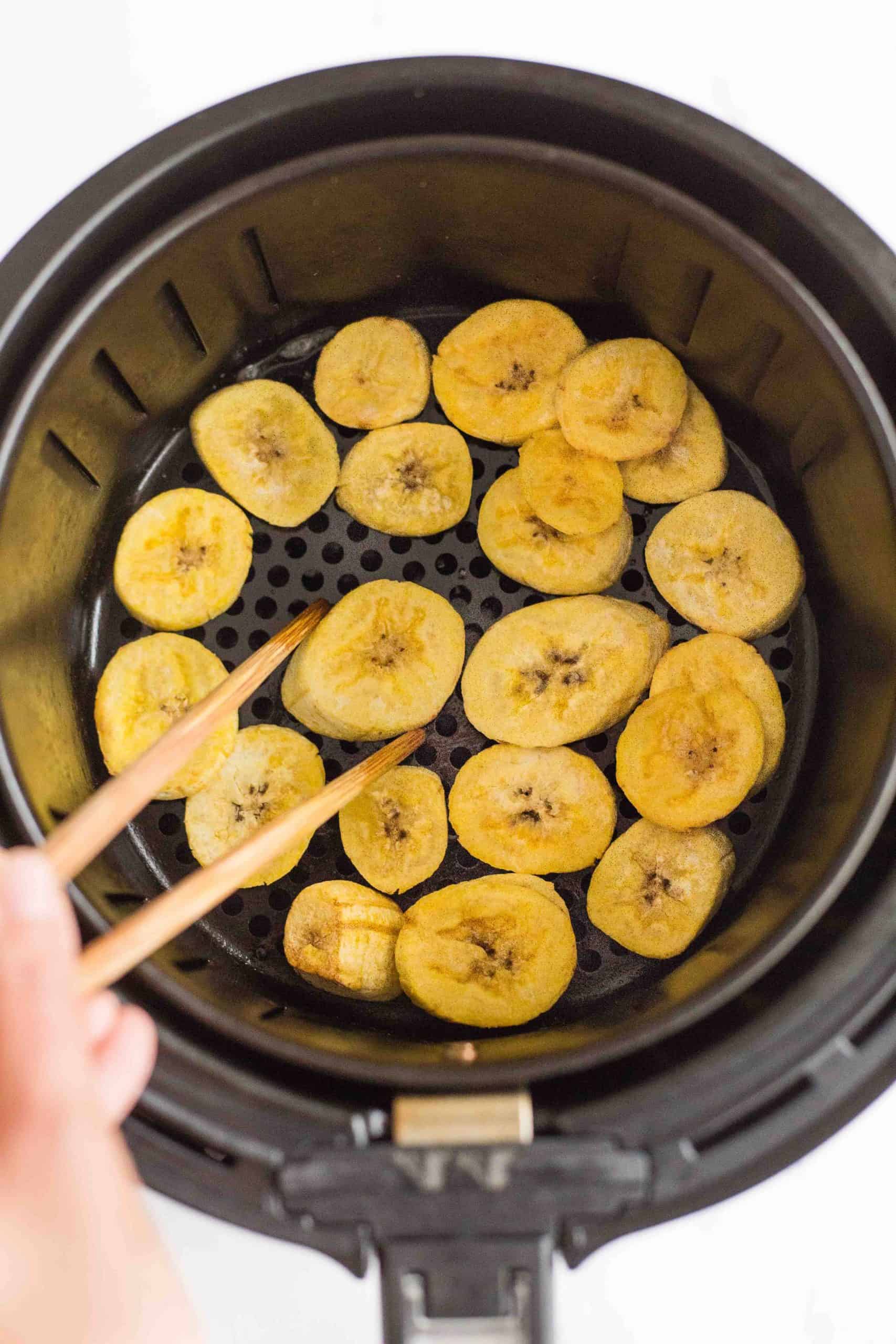 Using chopsticks to flip over plantain slices in air fryer basket.