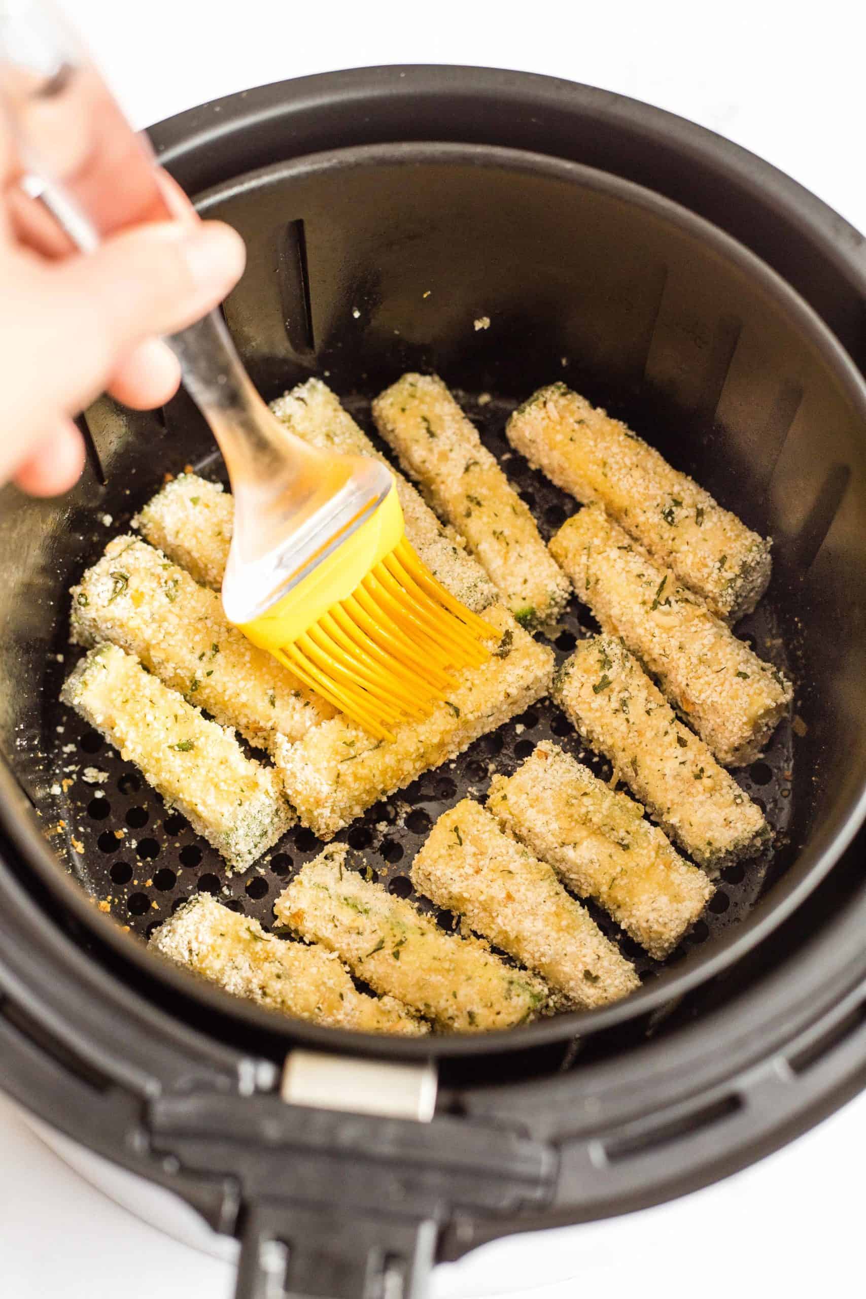 Brushing breaded zucchini sticks inside an air fryer with olive oil.