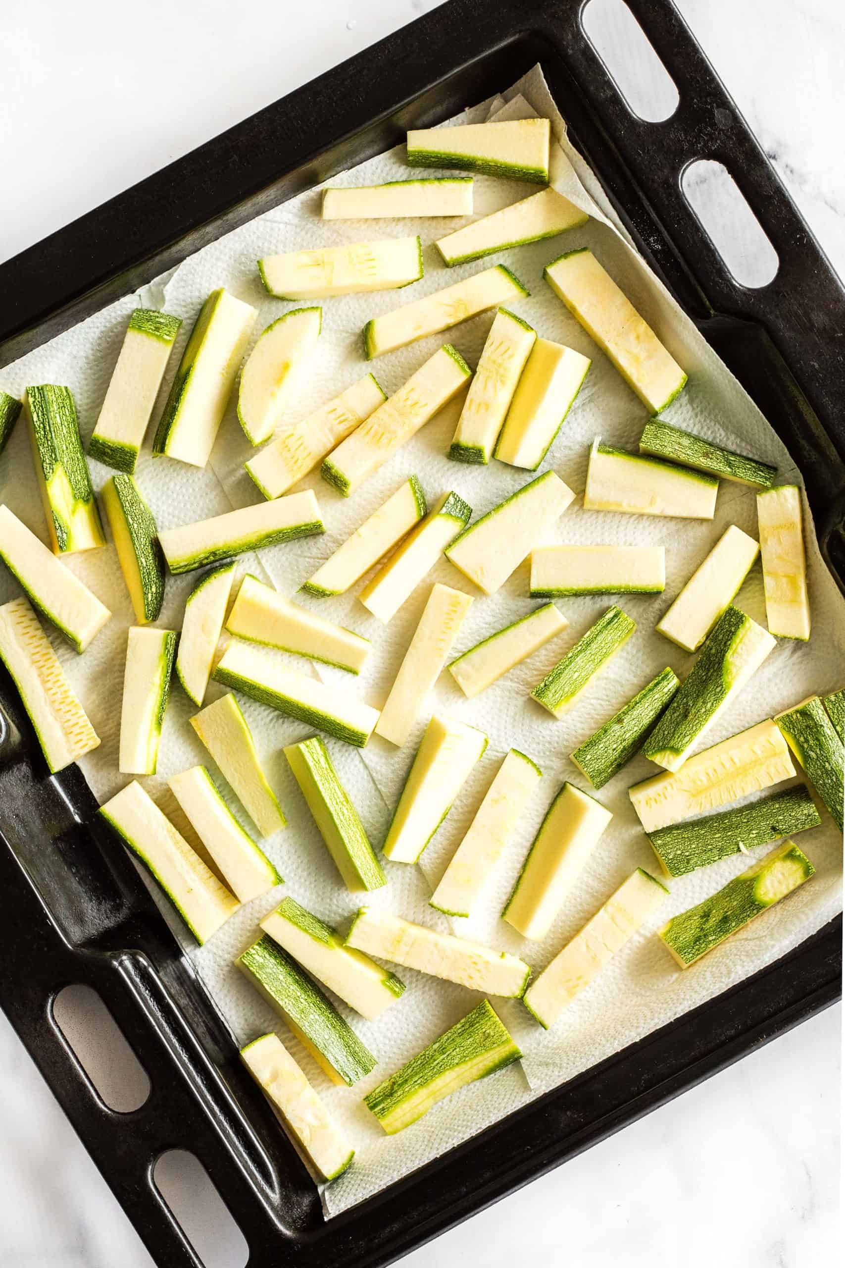 Cut zucchini sticks sitting on a paper towel-lined baking sheet.