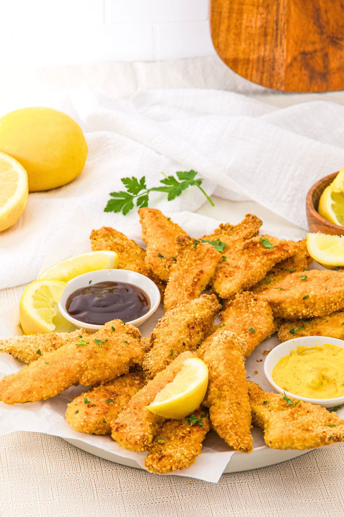 A plate of gluten-free chicken tenders with mustard and BBQ sauce and lemon wedges