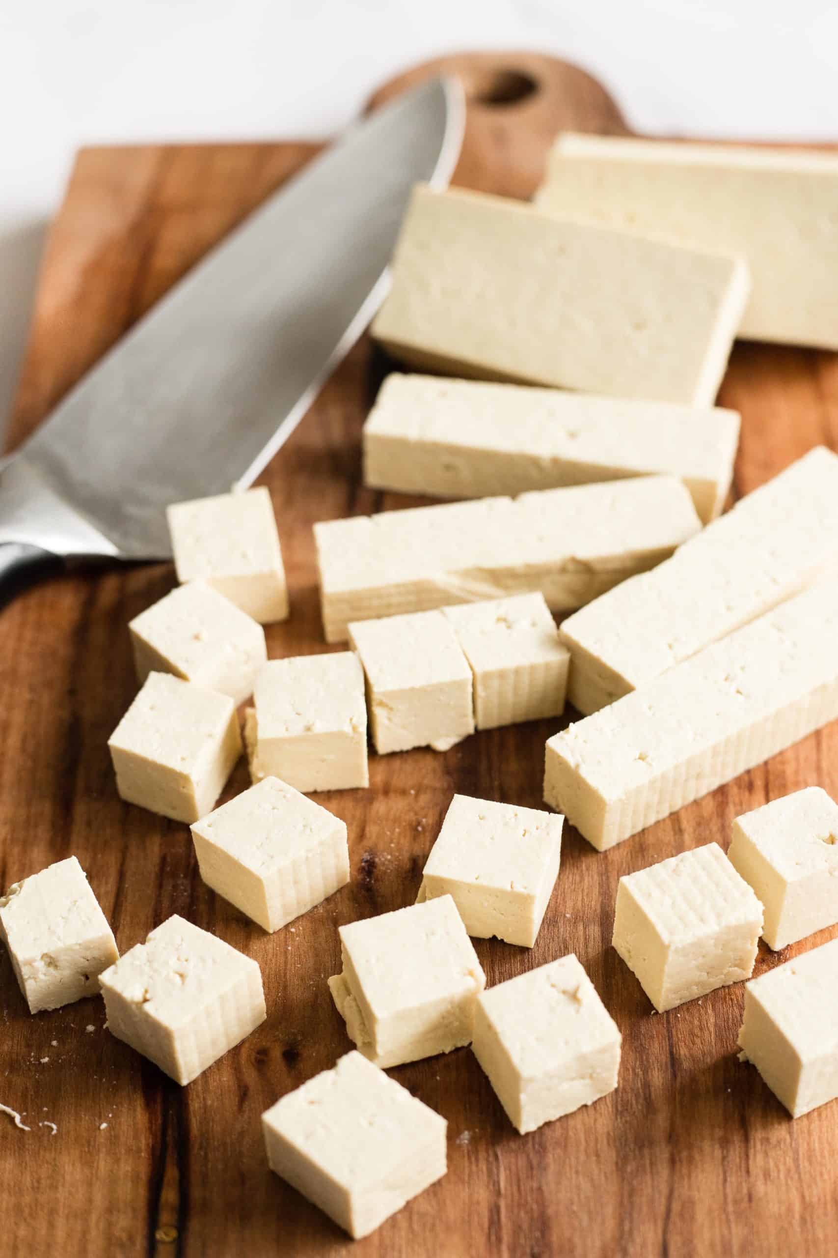 Tofu being cut into small cubes on a wooden chopping board.