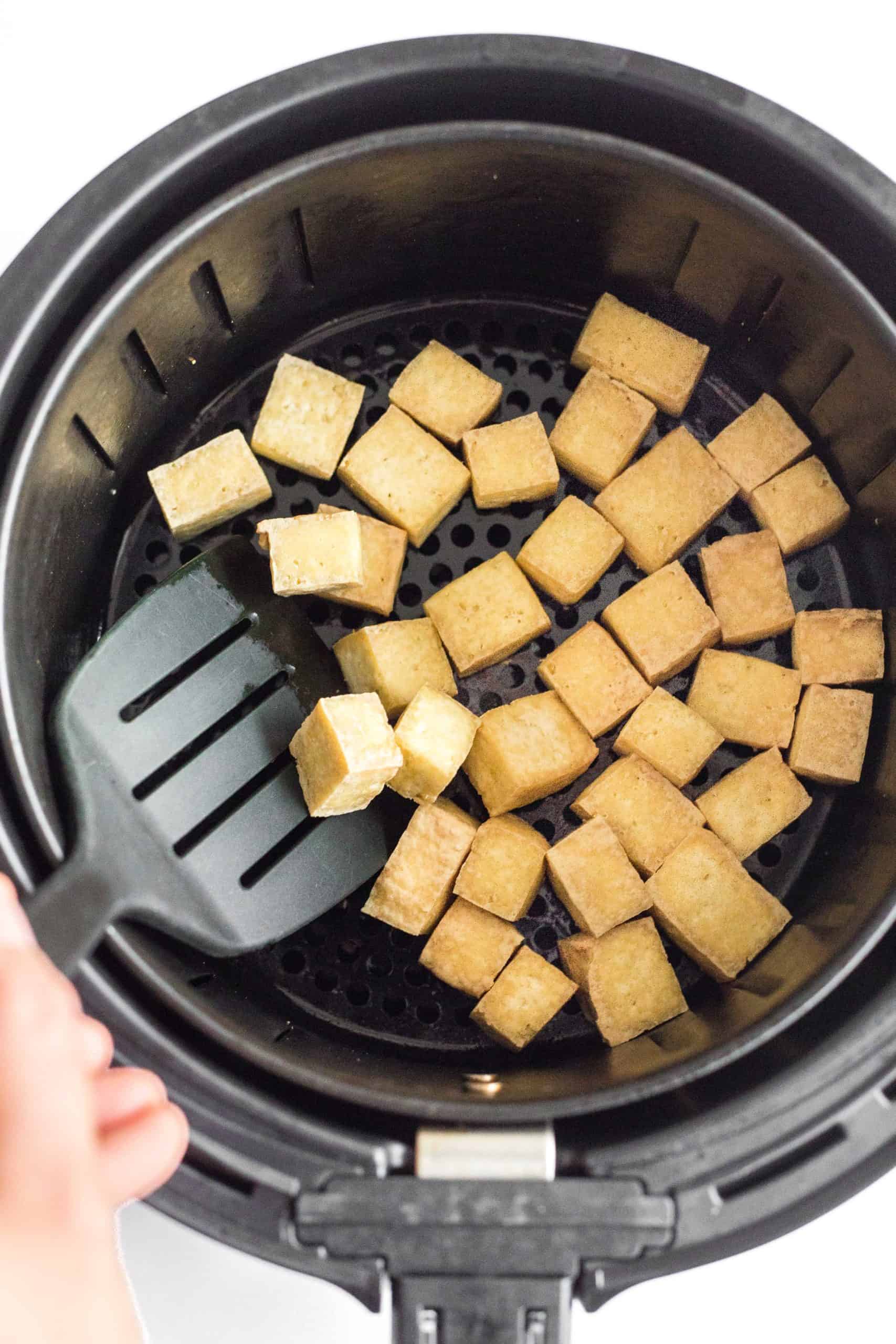 Hand tossing crispy tofu cubes in an air fryer basket after they are cooked.