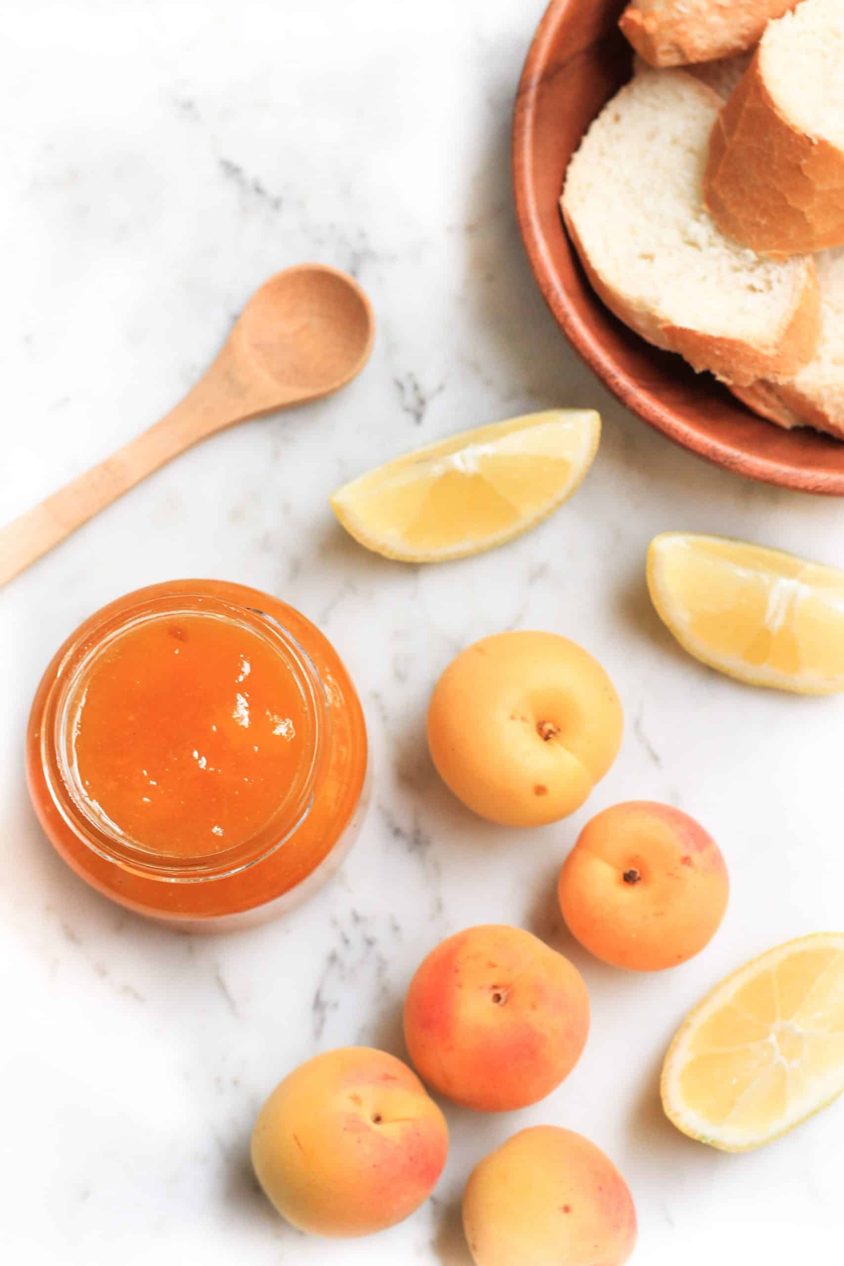 An open jar of apricot jelly, apricots, lemons, and bread on a marble board.
