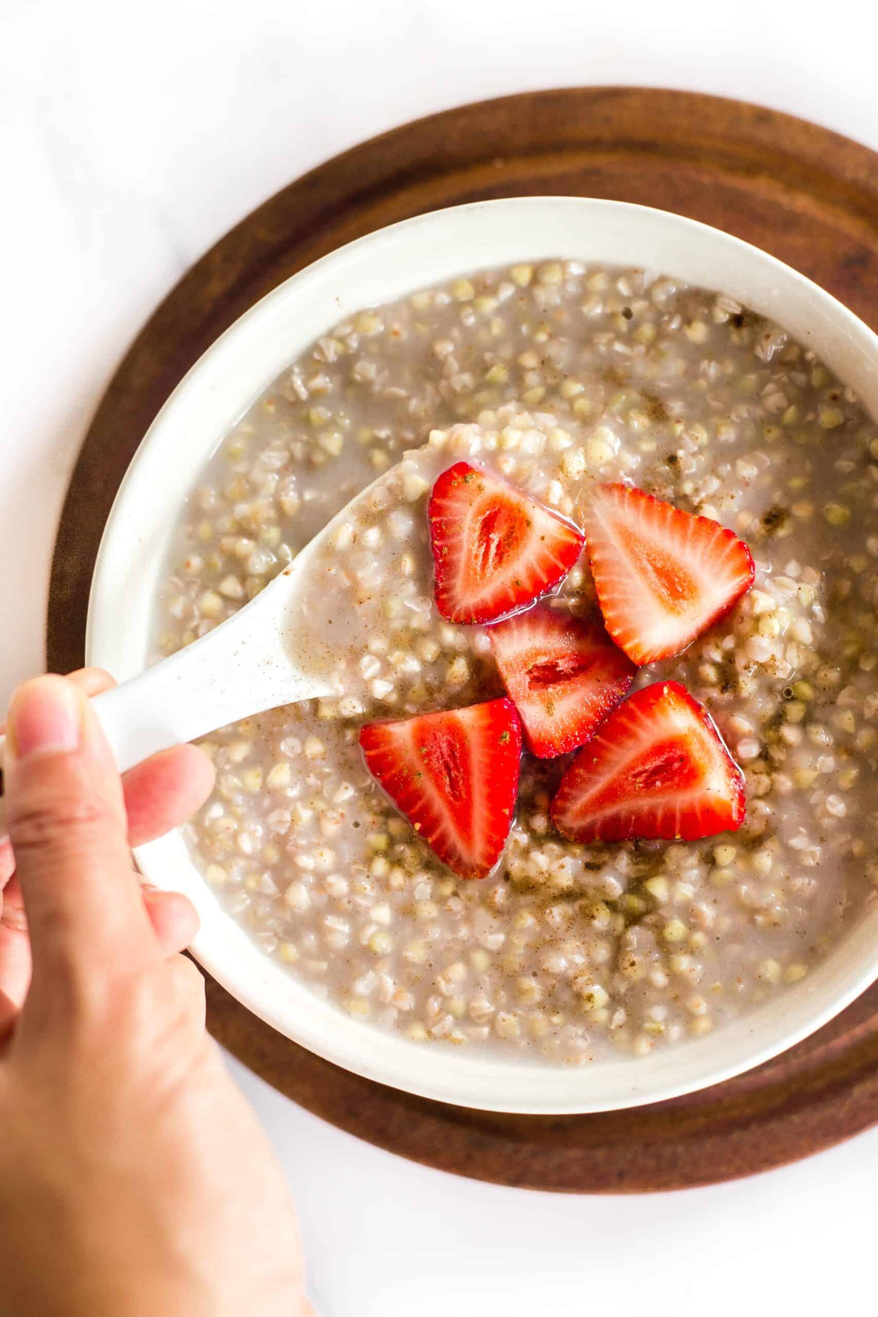 Hand holding a spoon in a bowl of buckwheat porridge with sliced strawberries.