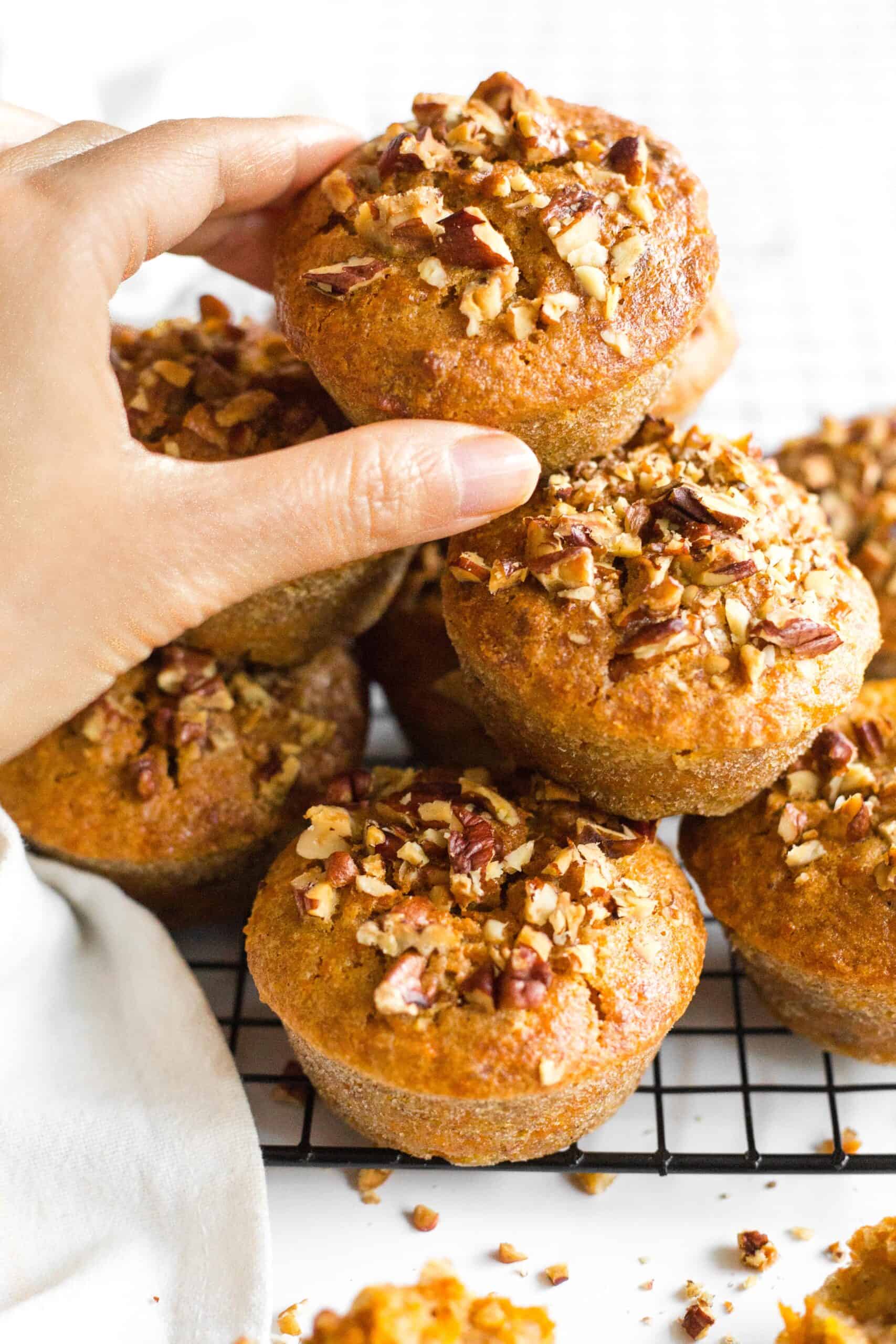 Hand reaching for a carrot muffin from a stack of gluten-free carrot cake muffins on a rack.