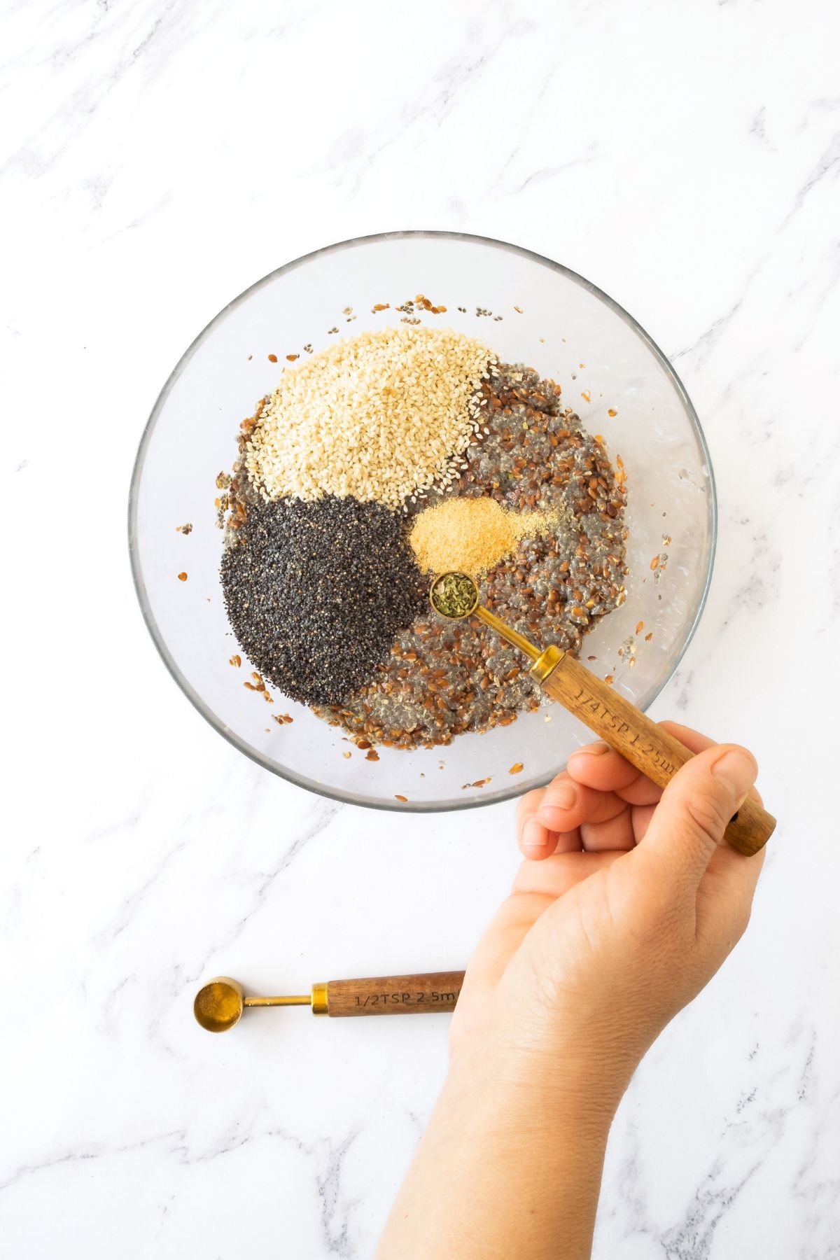 Adding dried oregano to a bowl of chia seeds, poppy seeds, garlic powder, sesame seeds.
