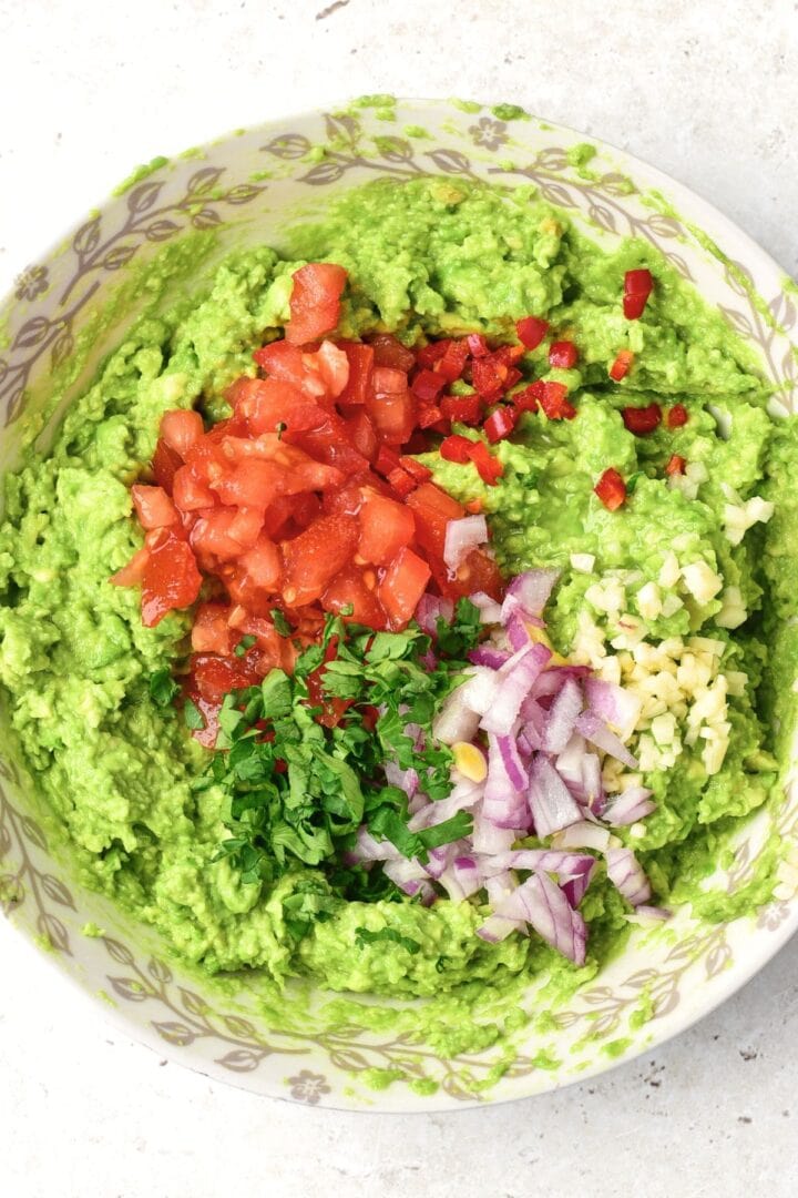 Chopped tomatoes, onions, cilantro, garlic on top of a bowl of mashed avocados