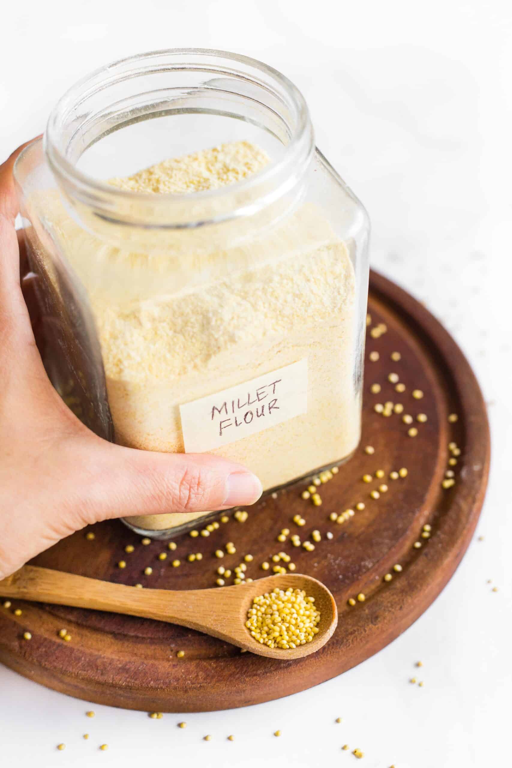 Holding a jar of homemade millet flour surrounded by millet seeds.