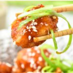 Chopsticks holding up a piece of fried chicken tossed in red sauce