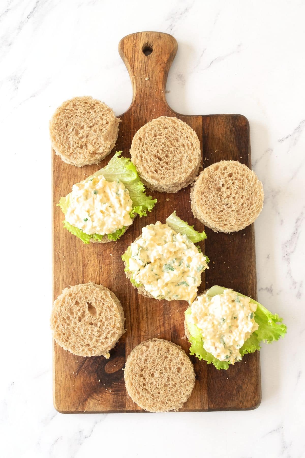 Preparing egg salad finger sandwiches on wooden board.
