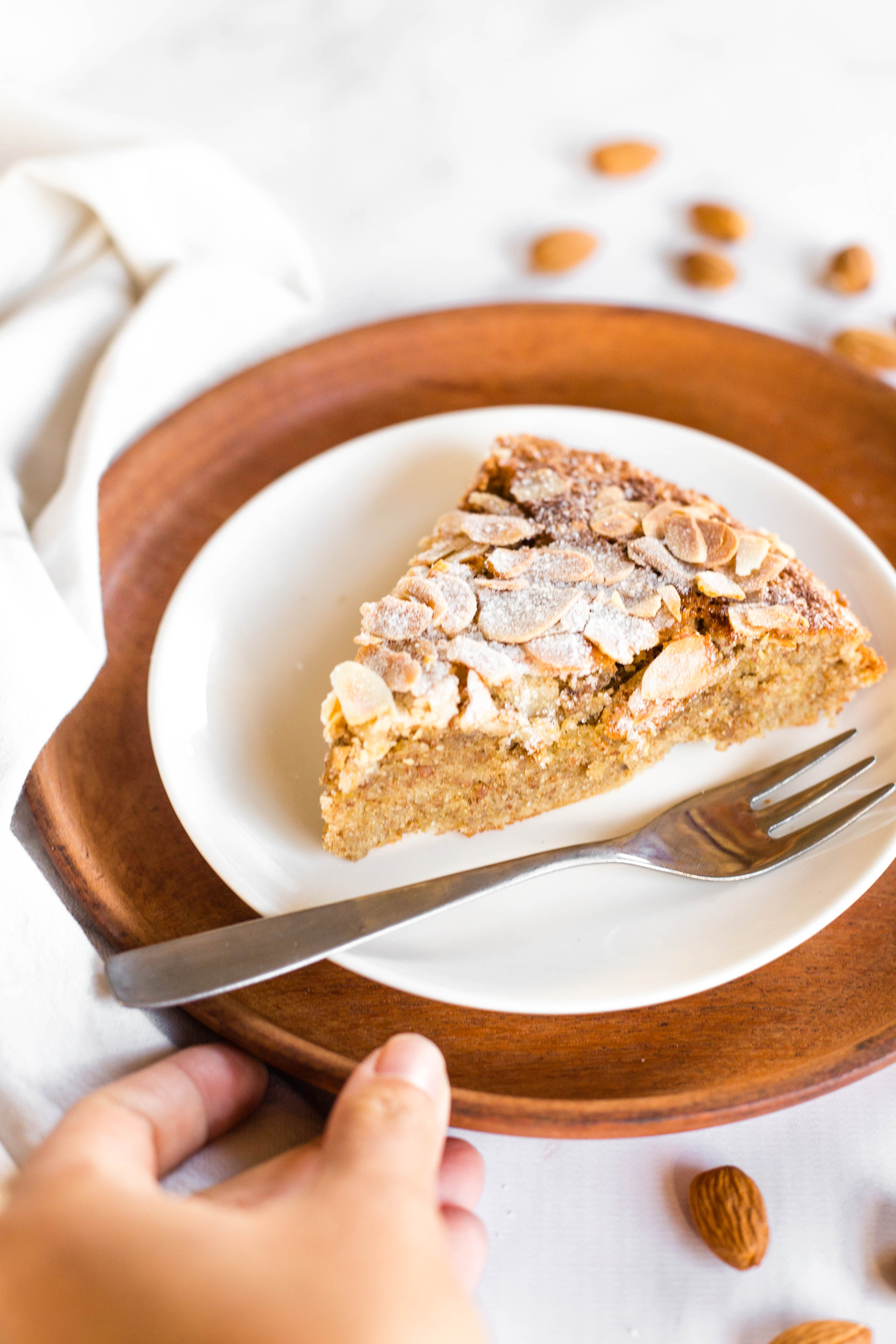 Reaching for a slice of gluten-free flourless almond cake on a white plate.