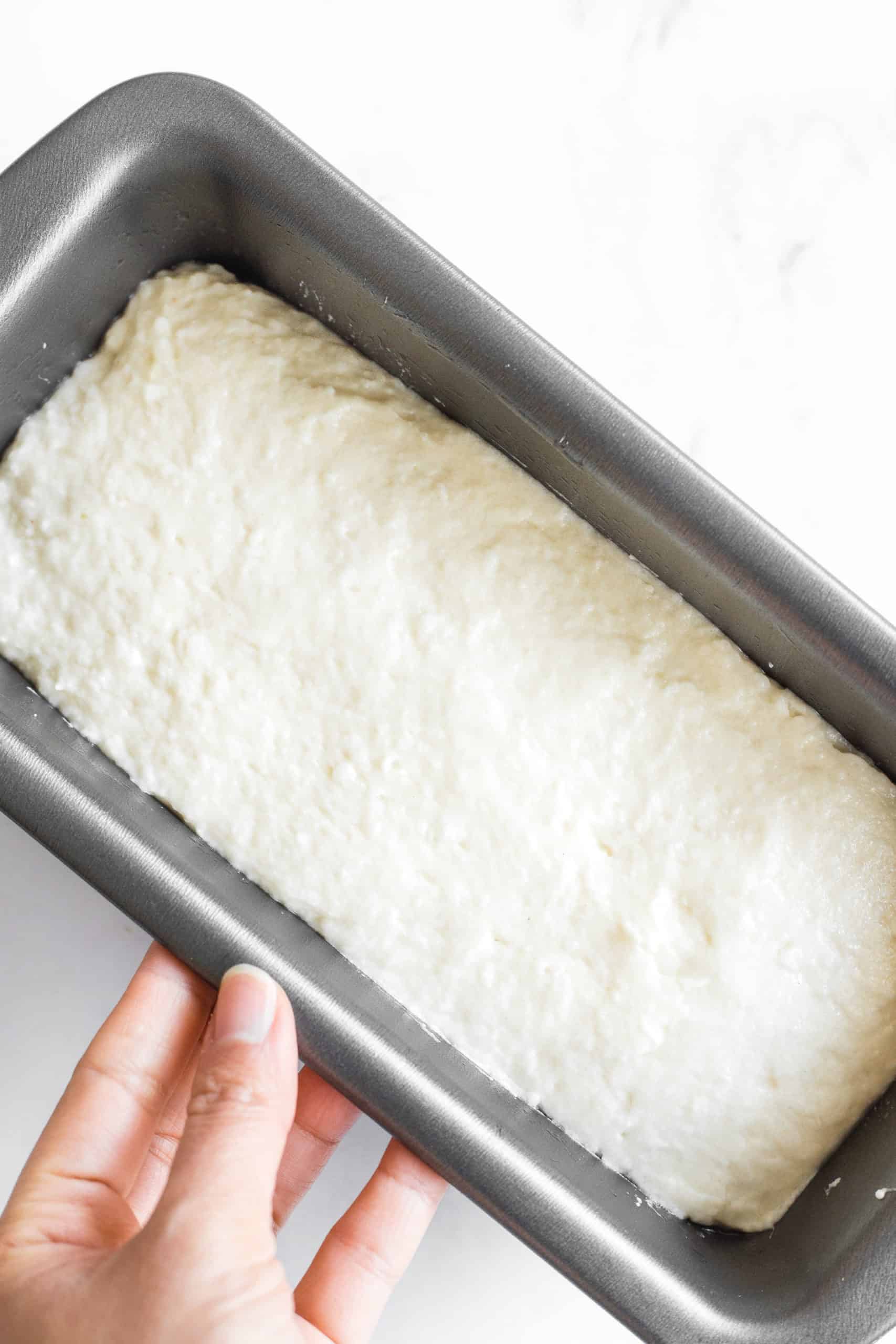 Hand holding a metal loaf pan with gluten-free bread dough before rising. 