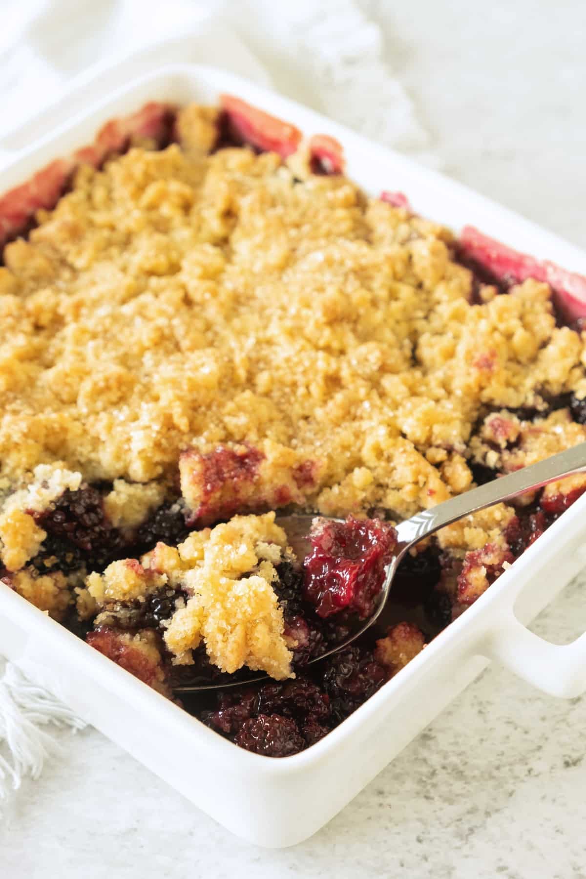 A spoon scooping into a blackberry cobbler in white casserole dish.