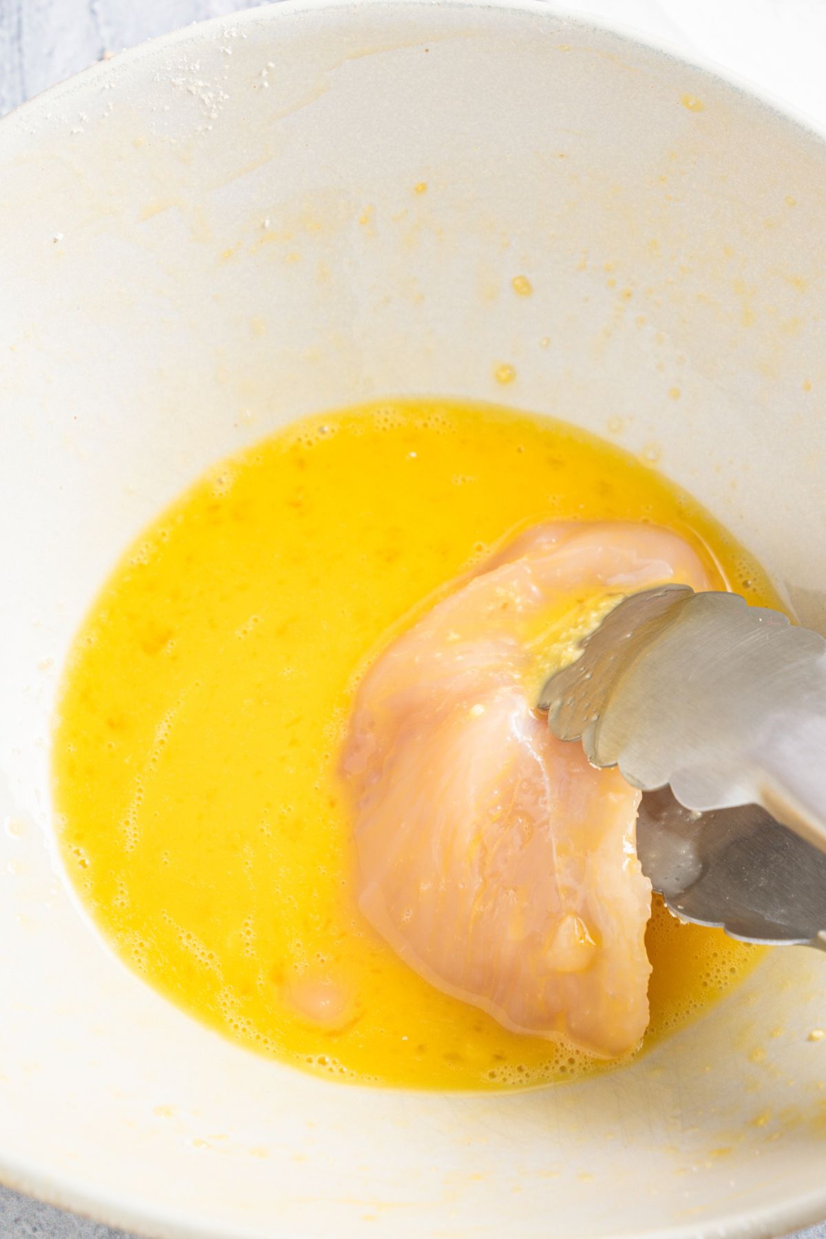 Using kitchen tongs to dip chicken fillet in beaten eggs.