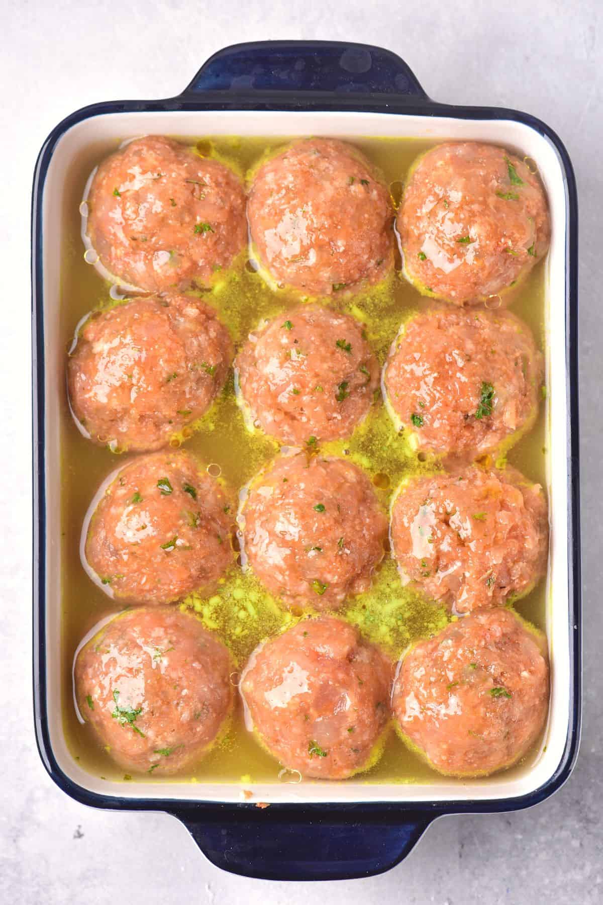 Uncooked chicken balls in broth in a baking dish.