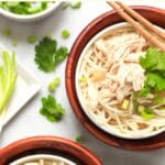 Two bowls of chicken pho surrounded by cilantro and lime