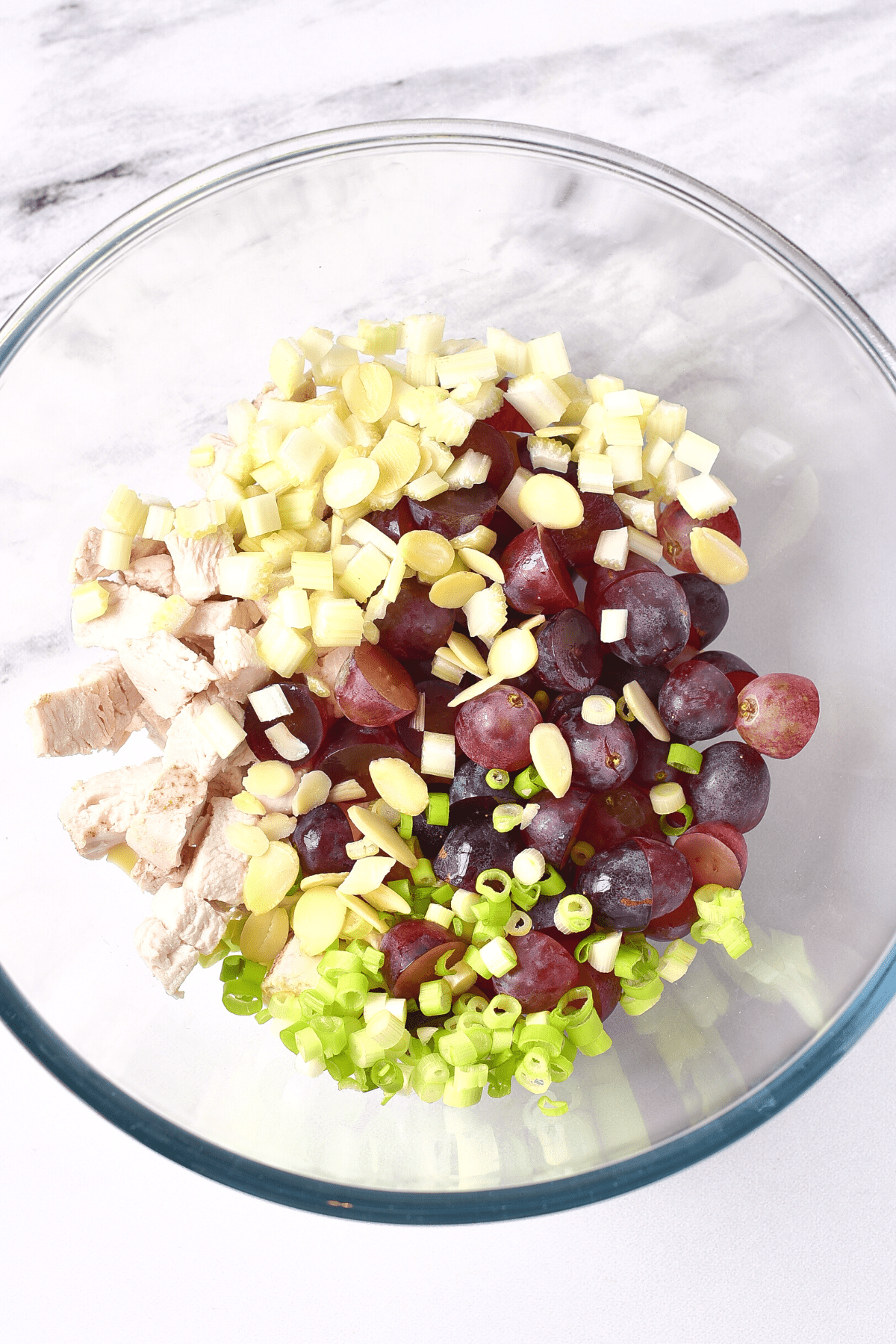 Chicken, grapes, celery, sliced almonds, and spring onions in a glass mixing bowl.
