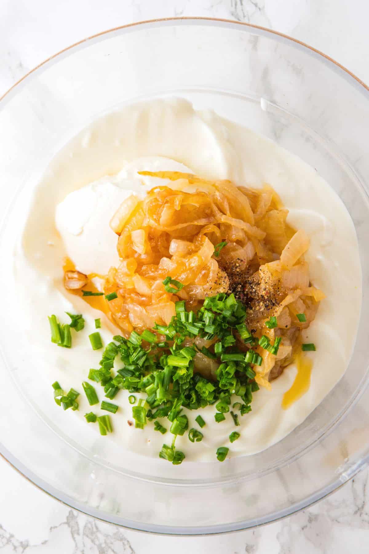 Caramelized onions, chopped chives, sour cream, and cheese cheese in glass bowl.
