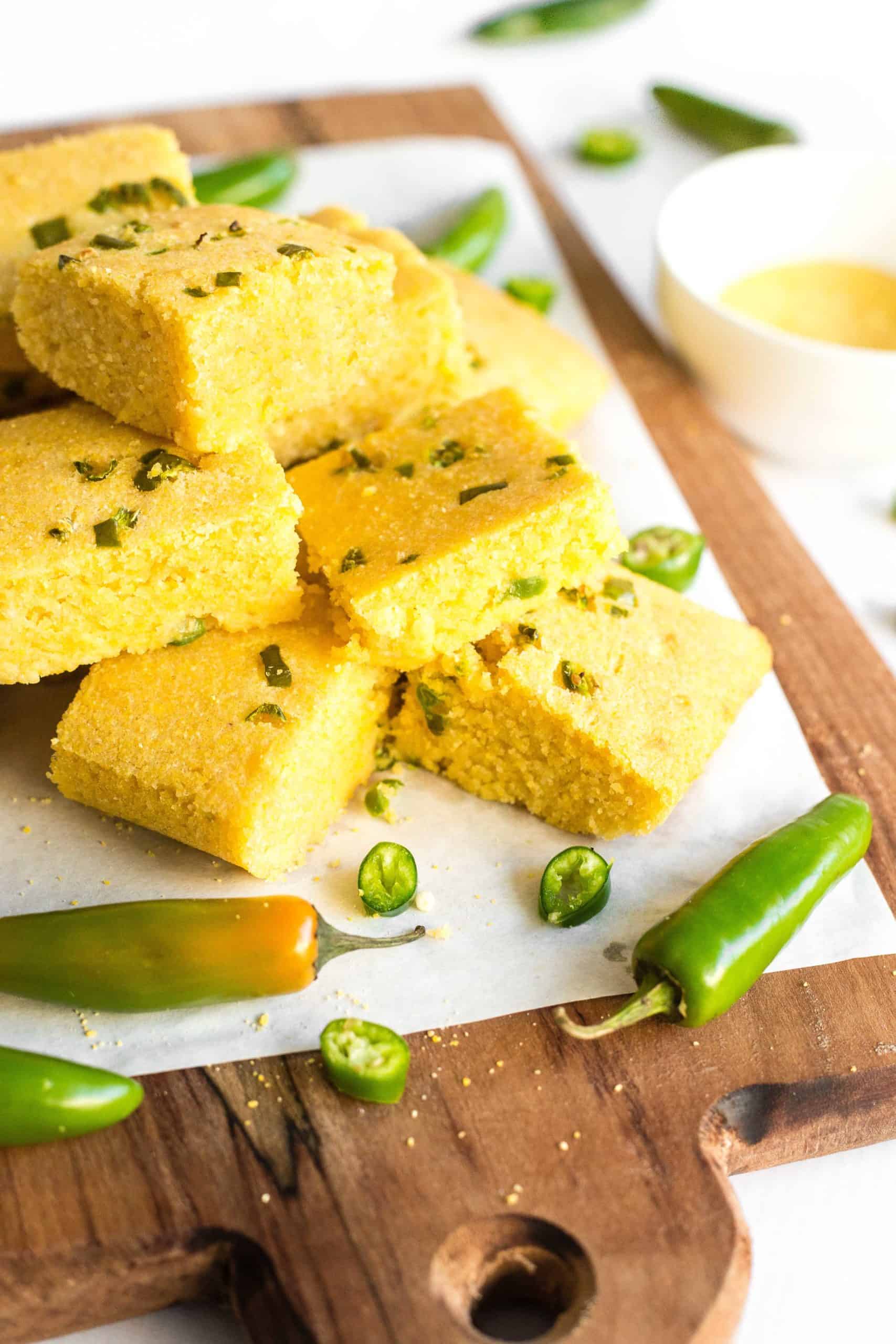 A stack of gluten-free jalapeño cornbread squares and fresh jalapeño peppers on a parchment-lined wooden board.