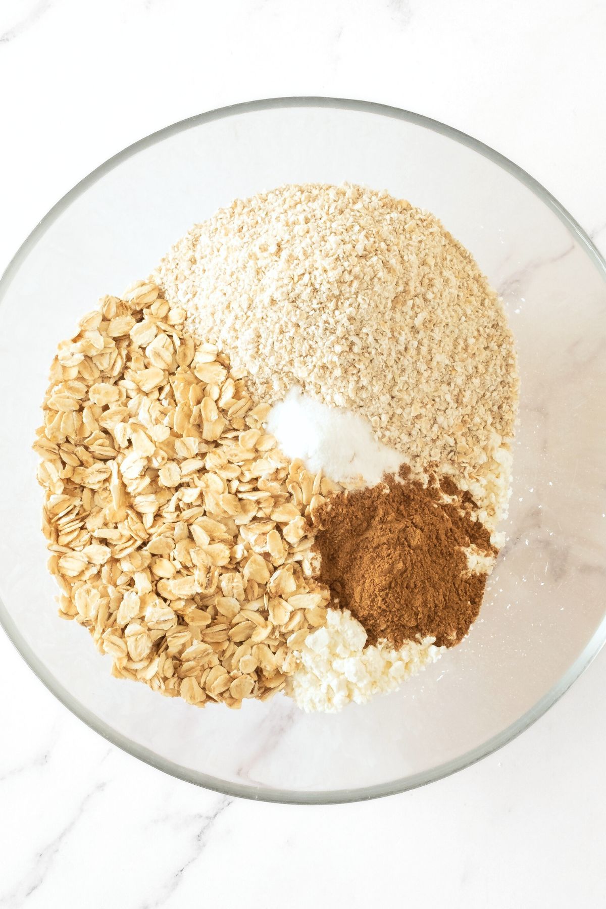 Rolled oats, gluten-free flour, ground cinnamon and baking powder in glass bowl.