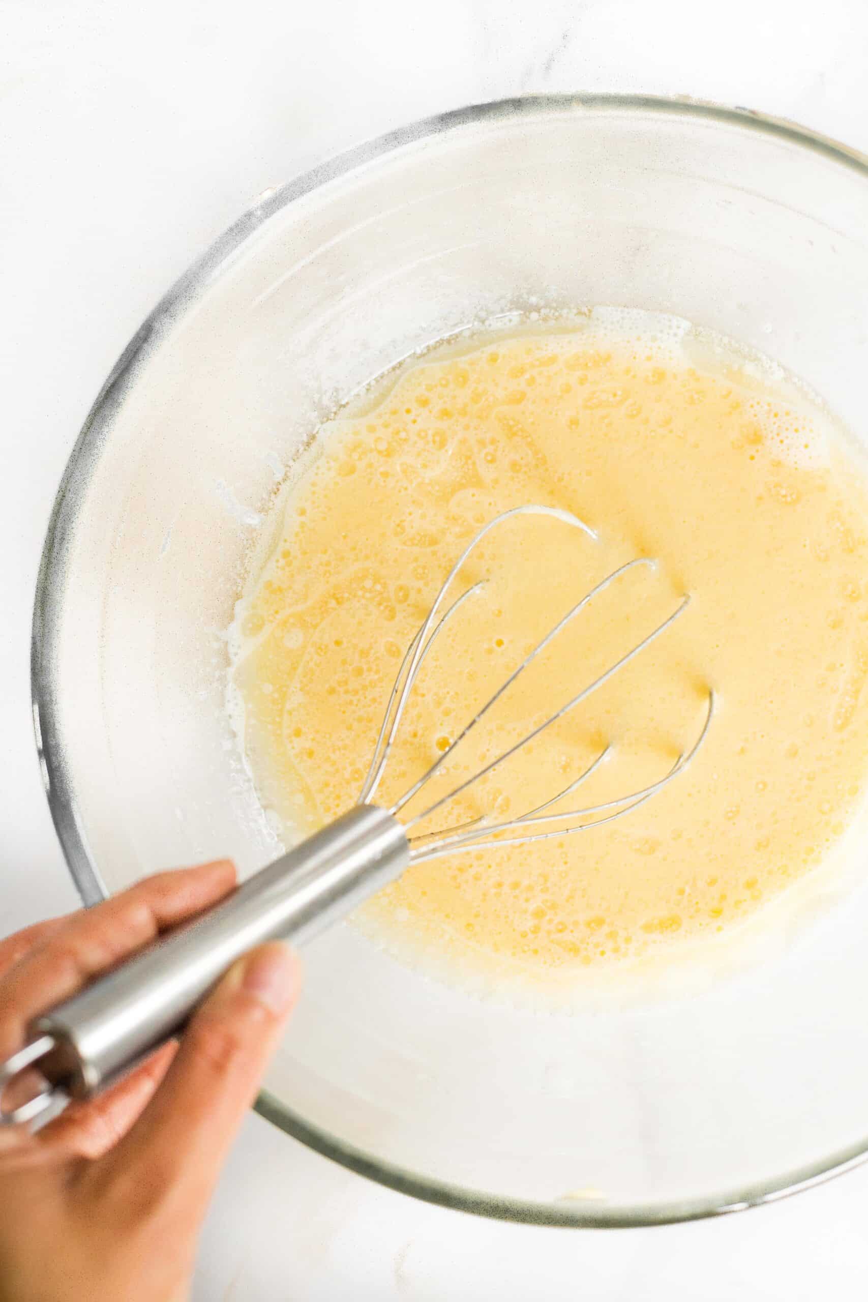 Whisking a pale yellow liquid mixture in a glass bowl.