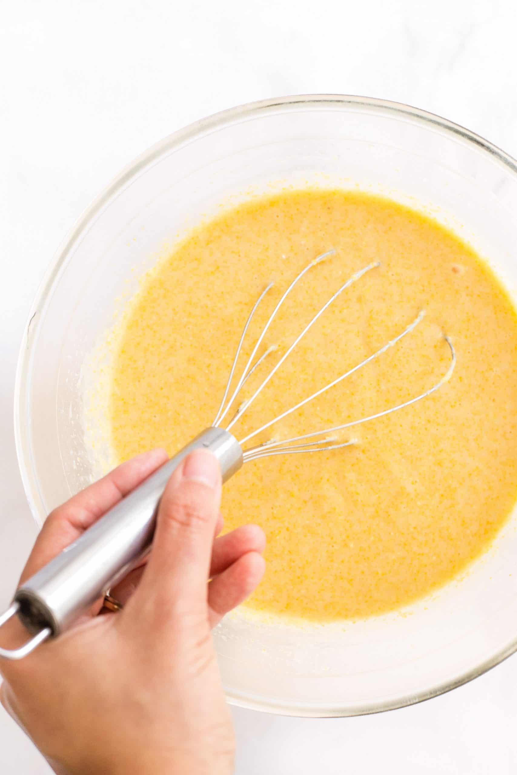 Hand whisking wet ingredients in a glass mixing bowl.