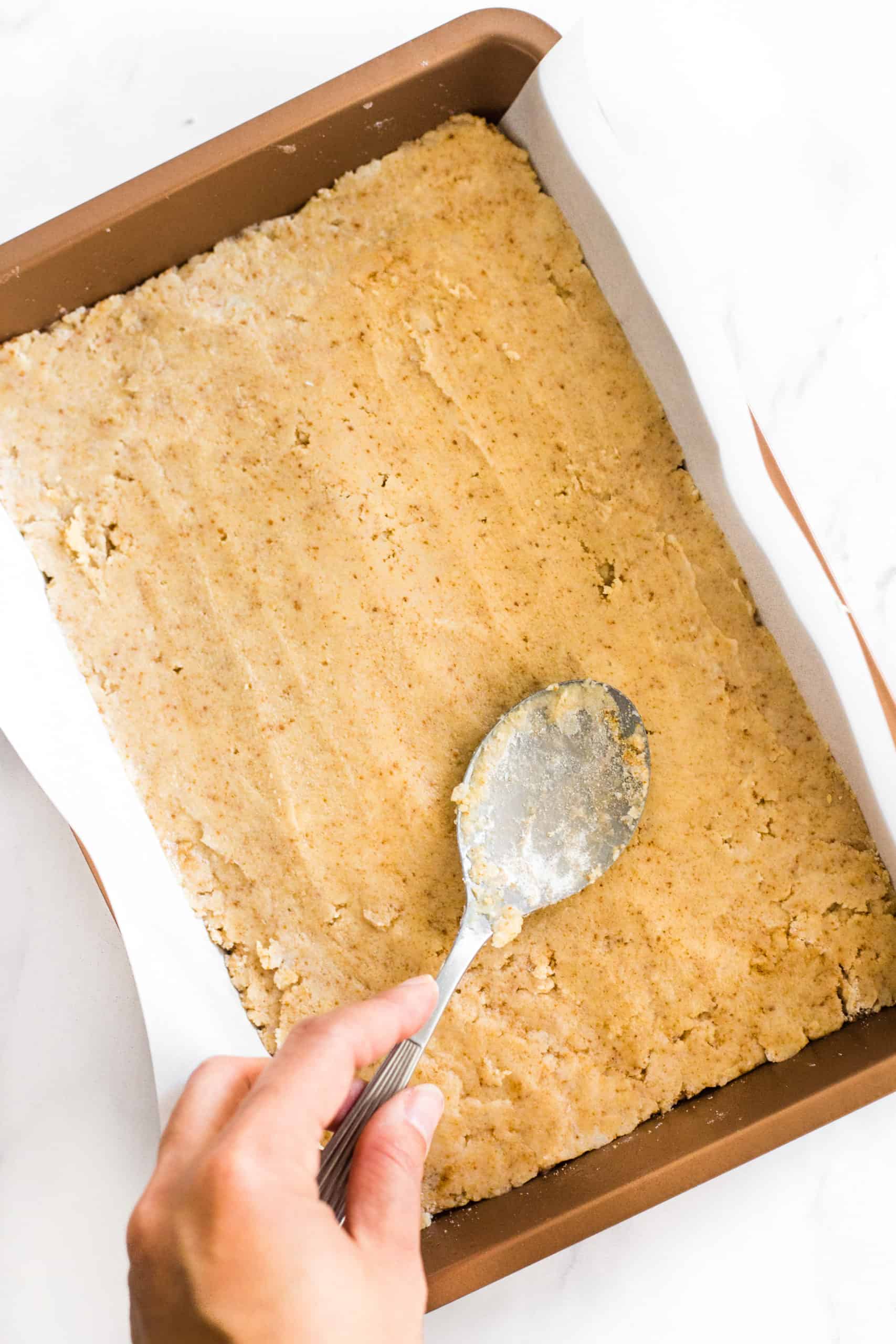 Crumb dough being pressed with the back of a spoon into a parchment-lined baking pan. 
