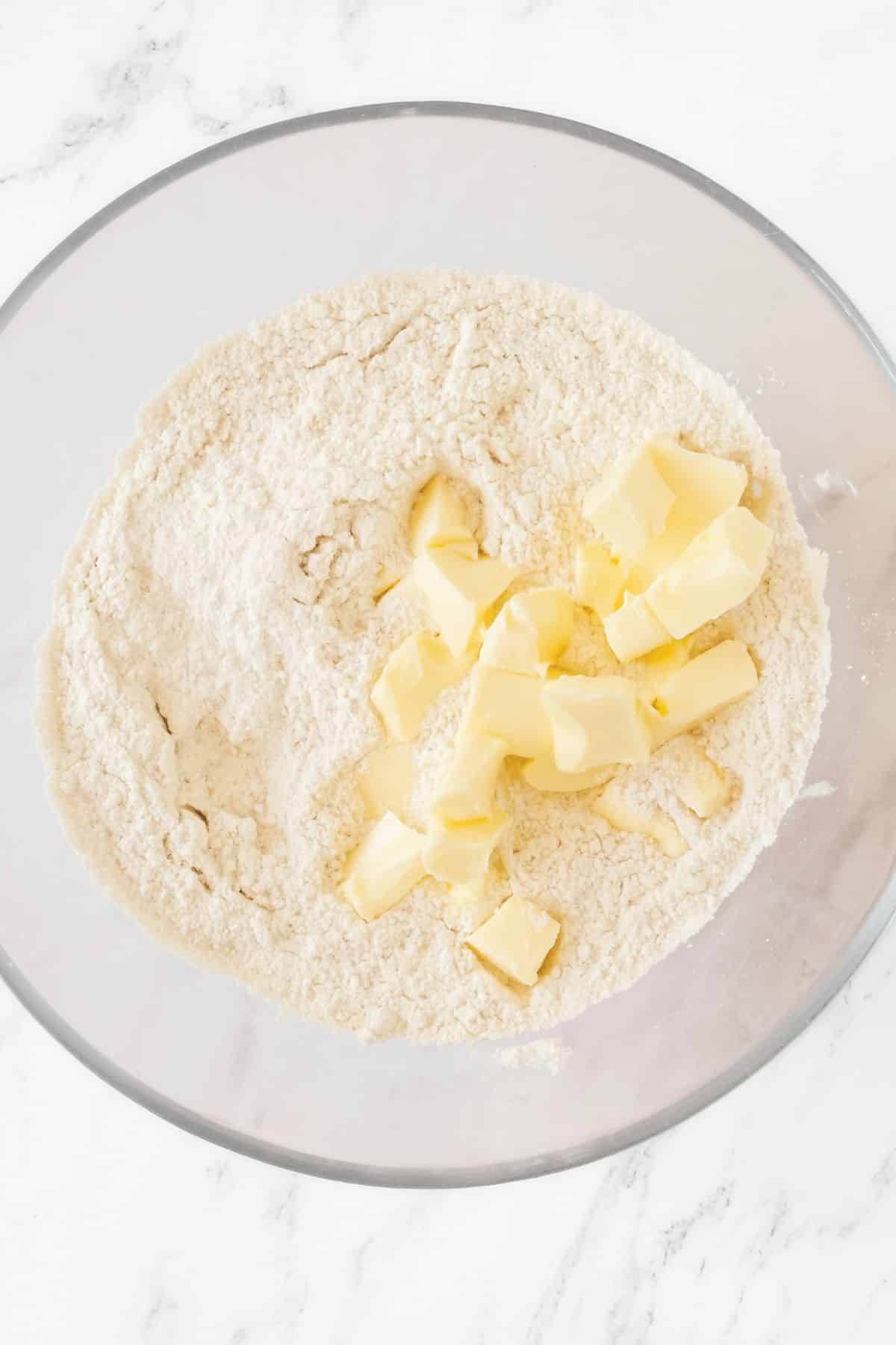 Cubed butter on top of flour mixture in glass mixing bowl.