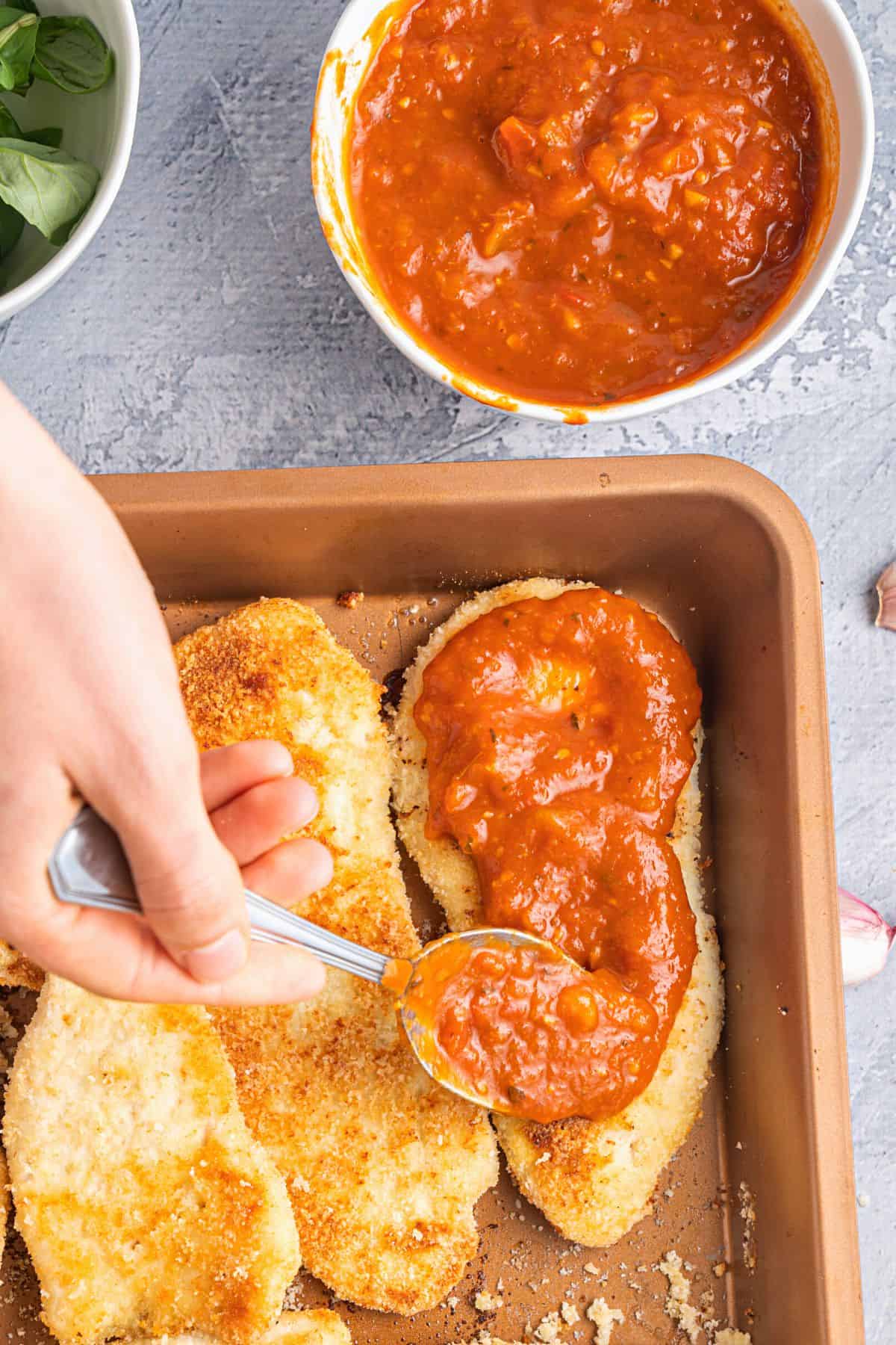 Spreading tomato sauce over breaded chicken cutlets.